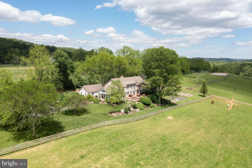Nestled in a private enclave of just five distinctive properties, Hideaway Farm is a 13+ acre gentleman’s estate offering both serenity and possibility. Currently configured for equestrian living, the land is equally suited for transformation—imagine a boutique vineyard, an heirloom orchard, or a curated retreat space. This is one of Upper Bucks County’s most coveted hidden locations: private, scenic, and truly exceptional.
Originally a humble ranch, the residence has been artfully reimagined into a transitional Craftsman-inspired home that blends country charm with understated elegance. Every room enjoys captivating views—some stretching for miles and even more striking when winter reveals the distant hills beyond the treetops. Step inside to a generous foyer with vaulted ceilings and flagstone floors—a true greeting room that sets the tone for the rest of the home. To one side, a glass-wrapped office opens to a large deck, ideal for client meetings or creative sessions in a space that feels as inspiring as it is professional. The dramatic living room features a soaring two-story wall of windows, clerestory rain-sensor windows, a brick wood-burning fireplace, and newly refinished pine floors. Open to the dining room, this central space is perfect for entertaining. In warmer months, step out to the 1,200 sf multi-tiered deck for al fresco dining with views of horses at pasture and the sun setting over rolling fields. The kitchen is both charming and functional, with granite countertops, glass-front cabinets, and a brick accent wall. A center island with propane cooktop and a full Bosch appliance suite—including double wall ovens, microwave, and warming drawer—make this a chef’s delight. The adjacent breakfast room includes a wet bar and beverage fridge and opens to a bluestone patio shaded by an awning—perfect for morning coffee with birdsong and the gentle splash of a nearby fountain. The main-level primary suite is a private haven with double-door entry, long-distance views, and its own balcony overlooking the pool. The spa-like en suite features a walk-in shower, jetted tub, dual granite vanities, and a large dressing room with custom storage. Upstairs, a lofted family room leads to three spacious, light-filled bedrooms with generous closets and stunning views. One bedroom offers a walk-in closet, cozy window seat, and private bath, while the other two share a hall with double-vanity bathroom. The walk-out lower level is an entertainer’s dream—complete with a wood-burning stove, new luxury vinyl tile flooring, billiards area, and a curved granite-topped bar with wine storage and three beverage refrigerators. There’s also an exercise room with rubber flooring, and a full bath and spa tub for post-workout relaxation. Just outside, a heated dark-bottom pool offers an idyllic spot to unwind amid the lush, verdant surroundings. For horse lovers, the property includes a 6-stall barn with Dutch doors, dry lot paddock, tack room, 300-bale hay loft, hot/cold wash stall, and 10 acres of clover and orchard grass pastures enclosed by 4-rail fencing. An outdoor riding ring completes the picture for serious or casual riders alike. Beyond the formal pastures lies a lightly wooded sanctuary—ideal for campouts, quiet fireside evenings, or off-the-grid escapes on your own back 40. Despite its peaceful seclusion, Hideaway Farm is conveniently close to Saucon Valley, Bethlehem, and Easton’s vibrant dining and shopping. With easy access to farmers markets, the Saucon Rail Trail, and just 90 minutes to NYC or Philadelphia, this is rural luxury with a cosmopolitan connection.