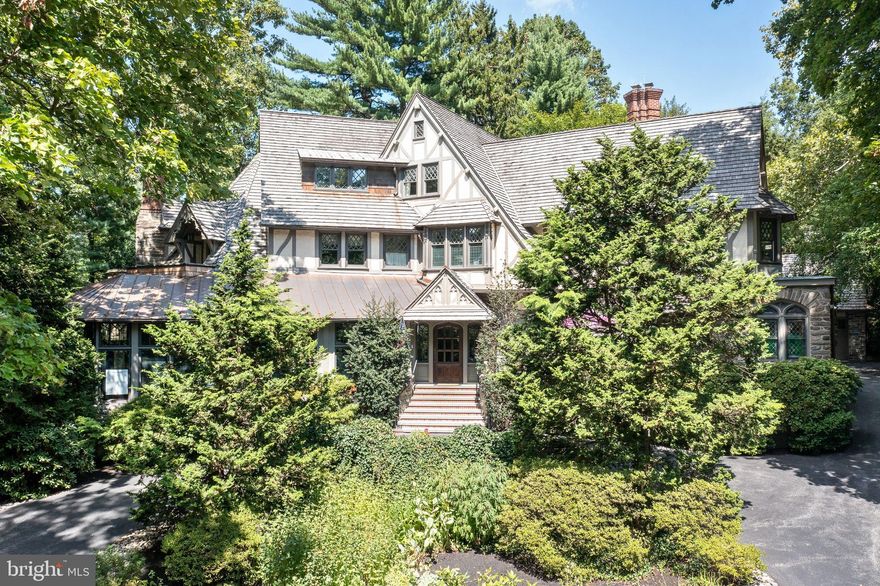 Follow the curving, tree-lined streets to your own private piece of history. Situated on a terraced southern slope in the South Wayne Historic District, surrounded  by old-growth oak, birch and Japanese maple, Orchard Hill is an outstanding, meticulously maintained example of Tudor Revival architecture within easy walking distance of downtown Wayne and the St. Davids train station.
Comprising 3 parcels on nearly 3 acres, including 414 Orchard Way and 502 Meadowbrook Circle, the offering includes the main manor home, a carriage house, 2 detached garages and a lighted sport court on a separately deeded parcel zoned for another residential structure if desired.
From cylindrical stone turrets, gabled cedar shake roof and leaded glass casement windows; to its oak flooring; quarter-sawn oak moldings, wainscoting and millwork; and 7 unique fireplaces, every inch of the main home is simply gorgeous. Beaded copper roofing over the sun porch and rear overhangs adds drama. Designed by the firm of noted architect William Lightfoot Price and built in 1899, the impressive stone-and-stucco manor combines old-world elegance and exceptional craftsmanship w/ contemporary amenities and upgraded utilities for maximum comfort and ease of living. The approximately 10,000-sf home features 16 rooms, including 5 bedrooms and 5 baths on its 3 above-ground floors.
Off the circular drive, a gabled oak entryway opens to a large center foyer. To the left, an arched doorway leads to a spacious living room with vaulted ceilings and original stained glass windows. The room’s focal point is an elaborately carved stone mantel featuring a pastoral tableau over 1 of 4 gas fireplaces on the first floor. Two sets of stained glass double doors provide access to an enclosed sunroom overlooking the flagstone patio and wooded grounds. And you’ll find a perfect spot for reading on a cushioned bench under the domed ceiling in the library/turret room, which features stained glass windows and curved bookshelves.
The living room also opens to the John Milner-designed eat-in kitchen, built by EB Mahoney, with Coventry cabinets and 2 dishwashers for holiday meals and large gatherings. Off the kitchen is a formal dining room w/  cozy alcove dining nook. The lower level has an 800-bottle climate-controlled wine cellar with soapstone countertops, Chicago brick and reclaimed French terracotta, Amish woodworking and hand-forged metalwork. A 2-story addition built in 2003 added a powder room, second office, mudroom and second-floor craft room/office adjacent to a large laundry room with linen closet.
Upstairs, escape to the primary bedroom suite, w/ 2 renovated en suite bathrooms, fireplace, custom closets, a window seat and both sunrise and sunset balconies. Two other second-floor bedrooms each have an adjacent full bath in the hallway, and the second floor is accessible from the foyer or kitchen. The third floor, with its architecturally unique rounded ceilings, has two additional bedrooms and a hall bath, as well as a sitting room/playroom and an air-conditioned attic with original built-in storage cabinetry. Second- and third-floor guest bedrooms also feature fireplaces, and some have generous window seats.
Throughout the home, main fixtures and utilities have been updated to include natural gas heat, central air and 400 amp electric, w/ a natural gas-powered back-up generator. It also boasts a water-softening system, wiring for electric vehicles in the 3-car main garage and commercial hi-speed WiFi throughout the compound.
Ideal for guests, in-laws or rental, a 2-story carriage house with slate roof has 2 upstairs bedrooms, plus a full bath on each level. It offers a remodeled kitchen and a spacious  living room with new flooring, built-ins and a beamed ceiling. It also houses controls for the heated saltwater pool with fountain, built-in spa and retractable cover. Carriage house has a 2-car garage w/ attic space.
Orchard Hill is in the highly desirable Radnor Schools.
