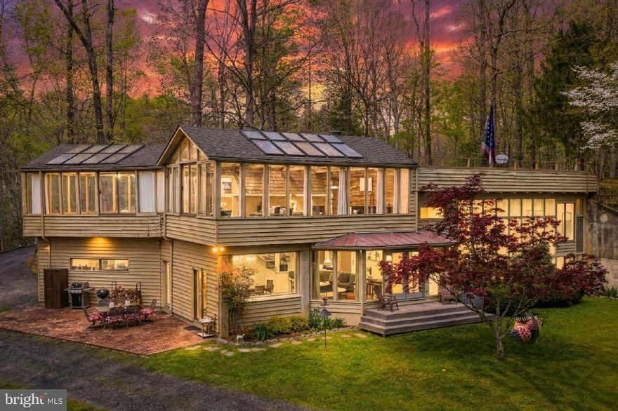 Tucked into a quiet stretch of wooded countryside, this totally architecturally distinctive retreat offers privacy, one-of-a-kind design, and a deep connection to the surrounding landscape. Located at 6251 Rokeby Lane in King George County, this 12.7-acre property includes a 3,758-square-foot residence evoking natural beauty and tranquil living, all without the constraints of an HOA! The approach is memorable. A beautiful gravel drive loops around the home, revealing a structure seamlessly built into the hillside and framed by hardwoods, ferns, and mossy ground cover. The front clearing opens to a peaceful expanse, while the remainder of the property remains wooded and serene, with a creek that feeds into the Rappahannock just beyond. A firepit, hammock area, woodshed, and thoughtfully placed trails with bridges and steps invite exploration, while the adjacent Dahlgren Historic Rail Trail adds another layer of outdoor adventure. The home itself elicits an immediate “Wow!” with its striking use of glass and natural materials. Designed with a passive solar concept by its original owner, an environmental engineer, the residence features expansive glass walls across both levels, balanced by concrete, wood, and original siding. A dramatic vertical glass element anchors the façade, while a newer front porch and curated gardens soften the architectural lines. The result is a home that captures light throughout the day and frames the surrounding landscape from nearly every vantage point. It is one-of-a-kind, folks!
Inside, the layout unfolds across a series of half levels, creating a sense of openness and flow reminiscent of Frank Lloyd Wright–inspired design. From the entry dining area, sightlines extend into multiple living spaces, each defined and connected. A signature glass panel sourced from Reagan National Airport separates the dining and living rooms, while terracotta tile flooring and exposed wood beams add warmth and texture. The kitchen has been fully updated with soapstone countertops, refreshed cabinetry, modern appliances, and dual sinks, blending with the home’s organic aesthetic. The bedroom layout offers comfort and flexibility. A guest wing includes two well-sized bedrooms with sliding glass doors that open to the front yard, along with an updated shared bath. The primary suite has been reimagined to include a renovated bath with a new shower, an expansive closet, and a separate study space that connects back to the main living area. Hardwood flooring runs throughout the bedrooms, complemented by wood ceilings and beams that carry the home’s cohesive design. Additional spaces enhance the home’s livability. A spacious sunroom on the upper level offers a greenhouse-like setting ideal for plants or relaxation, while the living room features a wood stove and generous proportions for gathering. The garage includes high ceilings, a utility sink, a large workbench, and direct access to a mudroom with a half bath. Recent system updates include a new well, HVAC, laundry, and updated flooring in the living room. Outdoor living is equally compelling, with a large brick patio, wooded trails, a shooting range platform, and a landscape that regularly welcomes wildlife, from songbirds to the occasional hawk or eagle. Positioned within 20 minutes of Dahlgren and Fredericksburg, and just minutes from local shopping and Fairview Beach, 6251 Rokeby Lane offers an incredible opportunity to live immersed in nature while remaining close to it all!