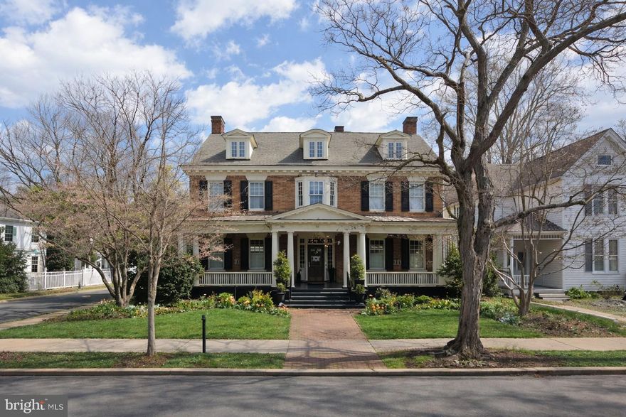 Are you looking for a distinguished downtown Fredericksburg residence that blends architectural elegance with modern luxury and unforgettable entertaining spaces? Positioned on a coveted corner lot within the Historic District, this exceptional 5-bedroom, 4 full and 3 half bath residence offers 5,239 square feet of refined interiors, bespoke craftsmanship, and a fully realized indoor-outdoor lifestyle—complete with a two-car driveway providing valuable off-street parking just steps from the city’s finest dining, shops, and riverfront. A gracious covered front porch with a richly detailed wood-paneled ceiling sets an impressive introduction, leading to a striking entry framed by transom windows and sidelights. Inside, the foyer reveals a sweeping curved staircase and integrated built-in coat closets, establishing architectural sophistication from the moment of arrival. The main level is defined by rich hardwood flooring, plantation shutters, oversized windows, and refined millwork. The formal dining room with chair rail and crown molding flows seamlessly into the gourmet kitchen, designed for both intimate gatherings and grand entertaining. An expansive island anchors the space, complemented by floor-to-ceiling custom cabinetry with soft-close drawers, display shelving, pull-out appliance storage, farmhouse sink, and a custom range hood highlighting the Wolf appliance suite with six-burner gas range, built-in oven, and microwave. A well-appointed butler’s pantry and discreet half bath enhance functionality, while a private office with built-ins and patio access adds versatility. The kitchen opens to a gracious family room with statement fireplace and custom shelving, while a formal living room with gas fireplace provides an additional refined gathering space. French doors reveal a sophisticated bar room featuring cork flooring, custom cabinetry, sink, ice maker, wine storage, arched windows, and backyard access—designed for elevated entertaining. Upstairs, the primary suite is introduced through glass French doors and includes a sitting room with bay window seating and storage, custom built-ins, and hardwood flooring. The spa-inspired bath offers dual vanities, walk-in shower with seating, separate water closet, and a custom walk-in closet with integrated laundry. Three additional bedrooms provide generous storage and architectural detail, complemented by a beautifully appointed hall bath. The third level presents a finished attic retreat with exposed beams, mini-split system, full bath, and flexible living space ideal for guests, studio, or private quarters. The partially finished lower level extends the home’s versatility with exposed brick, wet bar with wine refrigerator, half bath, exterior access, and a flexible guest or fitness suite complete with Murphy bed and full bath featuring a steam shower. Ample unfinished storage and expanded laundry capacity provide added convenience. The fenced rear grounds offer a brick patio and manicured turf framed by mature landscaping for privacy. The detached two-car garage has been transformed into an extraordinary all-season pavilion with fireplace, Wolf grill, quartzite countertops, Sub-Zero refrigeration, bar seating, hot tub, and half bath—an amenity rarely found within the Historic District. Don’t miss the opportunity to own one of downtown Fredericksburg’s most comprehensively appointed residences.