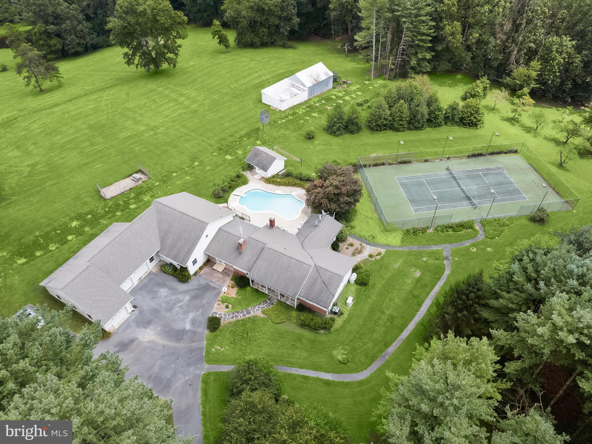 Discover the rare opportunity to own this exceptional six bedroom, four-and-a-half-bathroom home set on over 21 acres of natural beauty and seclusion. Surrounded by trees, the property offers a private and peaceful lifestyle with thoughtfully designed outdoor spaces including an in-ground pool, a full-size tennis court, and your very own pond. A long, tree-lined driveway leads to the professionally landscaped grounds that immediately evoke a sense of calm and retreat as you arrive. Inside, the front living room welcomes with the warmth of a wood-burning fireplace and rich hardwood floors that extend throughout much of the home. Just off the living room, the formal dining area connects to the sunroom that is an inviting space perfect for entertaining guests or simply enjoying a quiet afternoon. The eat-in kitchen features a breakfast nook for casual meals, along with ample cabinetry, generous counter space, and laundry. Off the kitchen, a cozy yet spacious family room provides another gathering space, complete with a wet bar and doors leading out to the pool area. A full bathroom with a walk-in shower is tucked nearby, ideal for guests or post-swim use. A few steps up, the expansive addition introduces the luxurious primary bedroom suite, designed with a dramatic vaulted ceiling and a private balcony that overlooks the surrounding grounds. The ensuite bathroom highlights a jetted tub, dual vanities, and a walk-in shower. A walk-in closet provides ample storage, and an adjoining bedroom adds flexibility for use as an office, nursery, or lounge. This wing of the home also includes another spacious bedroom and a versatile bonus room with direct access to the garage and outside, perfect for use as a mudroom, office, gym, or additional bedroom. On the opposite side of the house, a secondary bedroom suite includes a private full bathroom, while two more bedrooms and a shared hallway bathroom complete this level. Downstairs, the lower levels expand the space even further, with a recreation room, half bathroom, and abundant storage options to meet a variety of needs. Step outside to experience the true essence of this property. Whether lounging by the pool, playing a game of tennis or volleyball, or exploring the extensive wooded trails that wind past a flowing creek and a peaceful pond, this setting encourages connection with nature at every turn. Despite its quiet and secluded feel, the property remains just a short drive from major commuter routes and everyday amenities. This unique offering brings together space, privacy, and endless potential for both relaxation and recreation, truly an extraordinary place to call home. Other highlights include a barn for outdoor storage, orchards, a whole-house generator, a spring-fed water resource for the barn and pool, and two leased propane tanks. Additionally, the dual-zoned HVAC had two units replaced within the last two years. An adjacent 4.83 acre parcel may be purchased with this property. Some images have been virtually staged.
