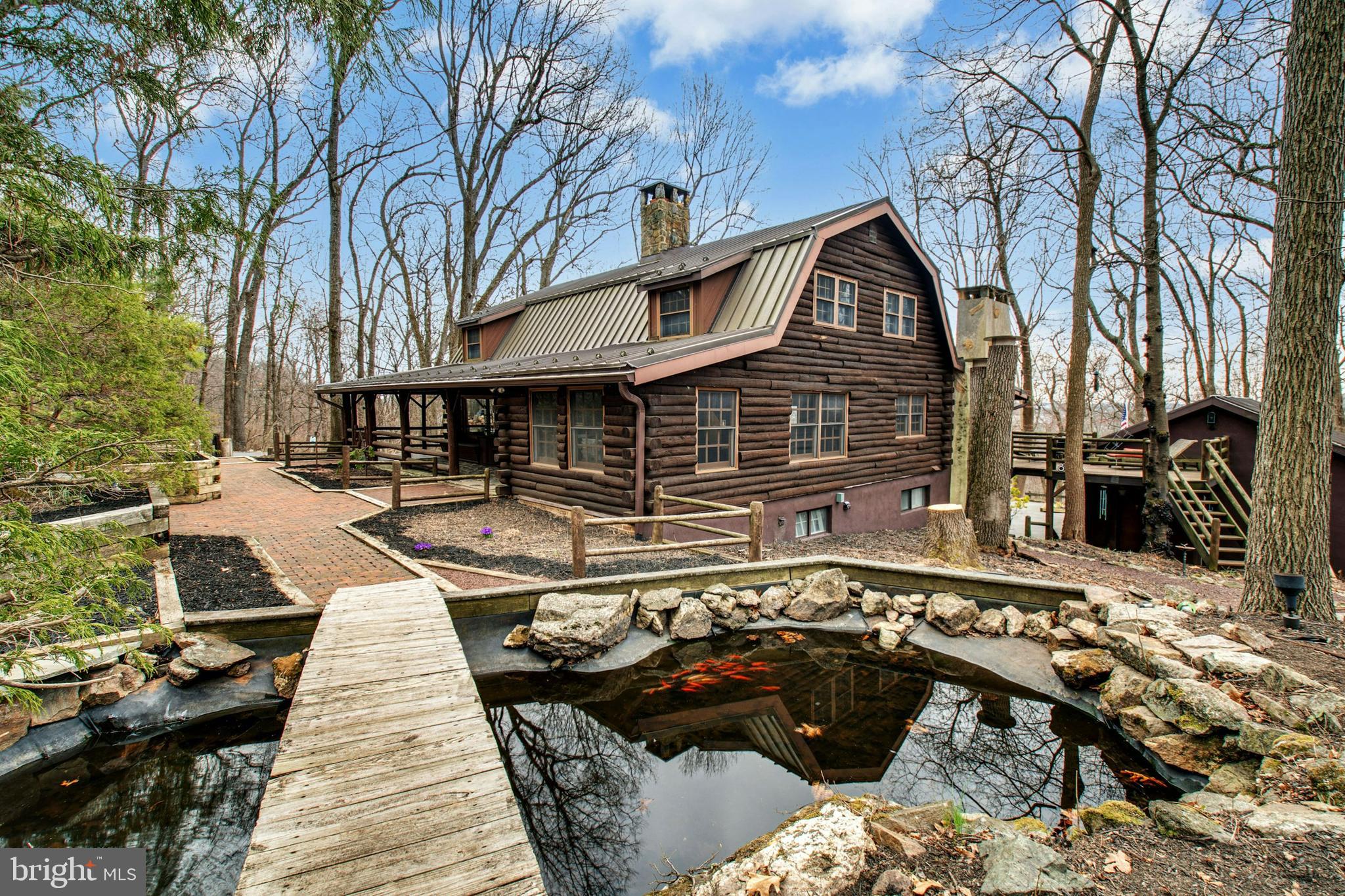 Crafted in a timeless tradition, Edgewood seamlessly blends modern innovations and comforts with a sophisticated rustic ambiance. Sheltered by tall trees, the 4.5+ acre parcel is set on a quiet country road in Lower Saucon Township.
A brick walkway leads guests and residents past inviting outdoor spaces including a koi pond spanned by a wooden bridge. From here, the stained glass front door opens to beamed ceilings, log walls, and exposed stone accents. The main level boasts a double sided stone fireplace open to both the living and dining rooms. A removable, custom built banquette between dining and kitchen areas can accommodate large parties or more intimate meals. The kitchen is light and bright with brushed aluminum cabinetry and stainless appliances, along with a walk-in pantry just around the corner. Office space and a half bath round out this level. Three spacious bedrooms and a full bath complete the second floor. The fully finished walkout lower level includes a wet bar, wood burning fireplace and full bath. An enormous covered wraparound deck includes an outdoor kitchen with grill and storage, and provides a front row seat to visiting deer, songbirds, and meticulously cultivated shade gardens.
Escape the city and experience the ambiance of a mountain retreat with close proximity to dining, shopping and cultural events in the Lehigh Valley and Doylestown.