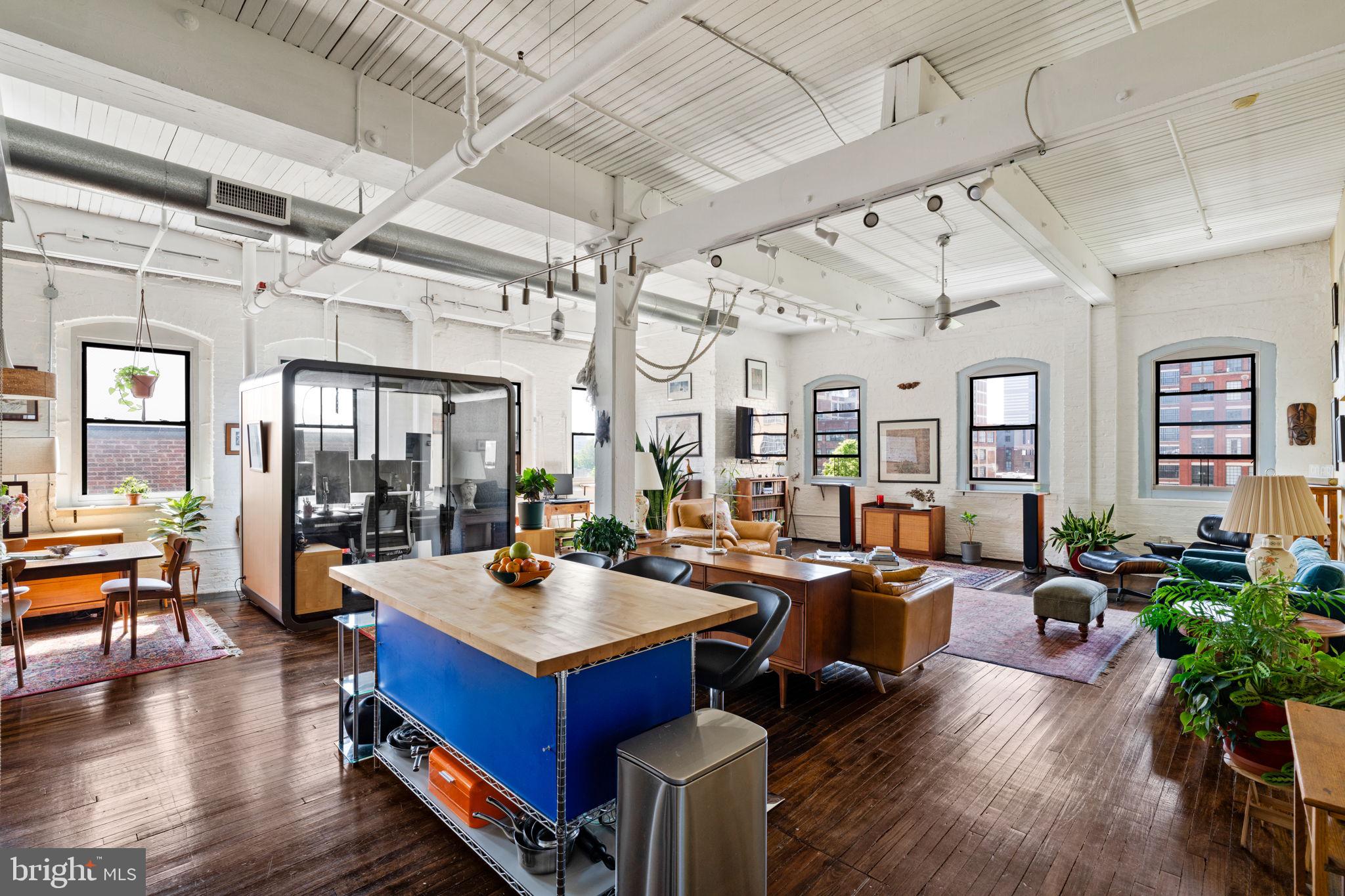 Magical and eclectic are two words that come to mind when entering this unique and sensorial space. Sunlight effortlessly floods through all eleven windows, creating a warm, cozy ambiance that delivers an instant feeling of zen.
Situated within the Callowhill neighborhood, in the Rebman Building, Unit 5G is a southeast-facing corner unit that offers direct views of The Rail Park and is a rarity on the market. Its 13-foot-high ceilings, storied original hardwood floors, and painted exposed brick walls fulfill its loft condo vibe requirements and most certainly assist in standing out from the monotony that strictens other condominium building concepts.
Essentially a large open living space, various layout options can be explored, including the addition of a second walled-off bedroom and bathroom, if desired. However, you may take inspiration from the owner's use of unique furniture pieces, artwork, lush houseplants, and many keepsakes, helping to delineate and make each room its own. You’ll even find wall and ceiling yoga ropes in the living room and office, which serve as mementos of the unit’s past as an Iyengar yoga studio. The owners agreed they should remain with the condo to commemorate the unit's provenance.
One-car heated and assigned garage parking with direct-to-floor elevator access is included in the sale, helping to establish this loft as a highly sought-after retreat amongst the surrounding gritty urban streetscape. Unit 5G will make you wonder, have you finally found your forever home?