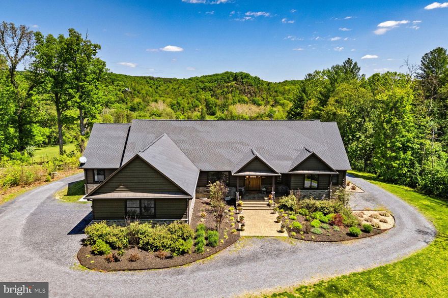 With over 1,000 feet of direct North Fork Shenandoah riverbank access, this 55-acre property offers a balanced mix of open pasture and mature woodland. In 2017, the current owners constructed a custom home designed to complement the setting, supported by additional structures including a renovated 19th-century bank barn guest house, a detached three-bay garage, and a separate tractor barn.
The residence was built to exacting standards using 2x6 framing, stone and HardiePlank siding, custom exterior doors, and locally milled hardwood flooring installed by Cochran’s of Berryville using lumber harvested from the property. The home offers 3,174 square feet of above-grade finished living space with three bedrooms, two full and two half baths, and a layout that places all primary living areas on the main level. An unfinished lower level provides approximately 850 square feet of storage with potential for future finishing, along with an additional one-car garage.
Antique doors sourced from around the world are incorporated throughout the home. The gourmet kitchen features a wood-topped center island and tumbled stone tile flooring, opening to a sitting area with wood-burning stove. This space flows directly to a deep, full-width, northeast-facing covered porch with elevated views of the river and surrounding mountains. Quality fixtures and fittings are used throughout, including William Morris wallpaper in select rooms.
A two-car side-load attached garage is complemented by a 40’ x 24’ detached three-bay garage suitable for equipment and storage. The 60’ x 30’ tractor barn, with an attached mechanicals room, offers flexible space for a variety of future uses.
Just below the porch, a tiered outdoor entertaining terrace includes an expansive concrete patio, integrated stone seating, landscaped beds, and a substantial wood-burning masonry fireplace set against a private wooded backdrop.
The circa 1830 bank barn, served by its own well and septic system, has been converted into guest accommodations featuring a kitchen, raised stage, sleeping quarters, one full and one half bathroom, cathedral beamed ceilings, and a second floor mezzanine/gallery, spanning much of the open interior.
A private riverfront property with acreage, multiple structures, and broad scenic views, located just over an hour from Washington, D.C.