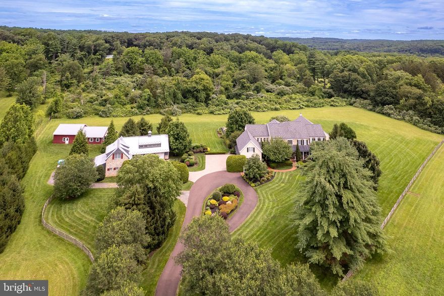 A winding driveway carves through almost ten acres of verdant scenery, arriving at a Bucks County Fieldstone home that feels plucked from a dreamscape. This custom-built beauty tucked quietly in the Solebury countryside, blends pastoral charm with refined grandeur offering a true sanctuary for the nature-loving aesthete. Inside, craftsmanship reigns supreme. Soaring ceilings on the main level pair with Brazilian cherry floors and extravagant millwork to set a tone of understated luxury. The two-story foyer, clad in marble and anchored by a sweeping staircase, makes a theatrical first impression. The gourmet kitchen is outfitted with soapstone countertops and Subzero and Viking appliances and warmed by a stone fireplace in the adjacent breakfast nook. A light-filled sunroom connects seamlessly to the great room, offering year-round views of the gardens and waterfall-fed pond. A first-floor guest suite and dedicated home office are tucked into a private wing. Upstairs, four en suite bedrooms extend hospitality and comfort, none more indulgent than the light-filled primary suite complete with a private sitting area, spa-inspired bath, and expansive walk-in closets worthy of a designer’s eye. An Adirondack-style indoor pool and spa wrapped in timber-lined cathedral ceilings and anchored by a wood-burning fireplace offers four-season relaxation. Auto enthusiasts will appreciate the eleven deep garage bays with epoxy flooring beneath a lovely carriage house. A heated pole barn with groundskeeping storage spaces offers endless possibilities for recreation. And when a night on the town calls, the galleries, theater, and dining of New Hope are only minutes away.