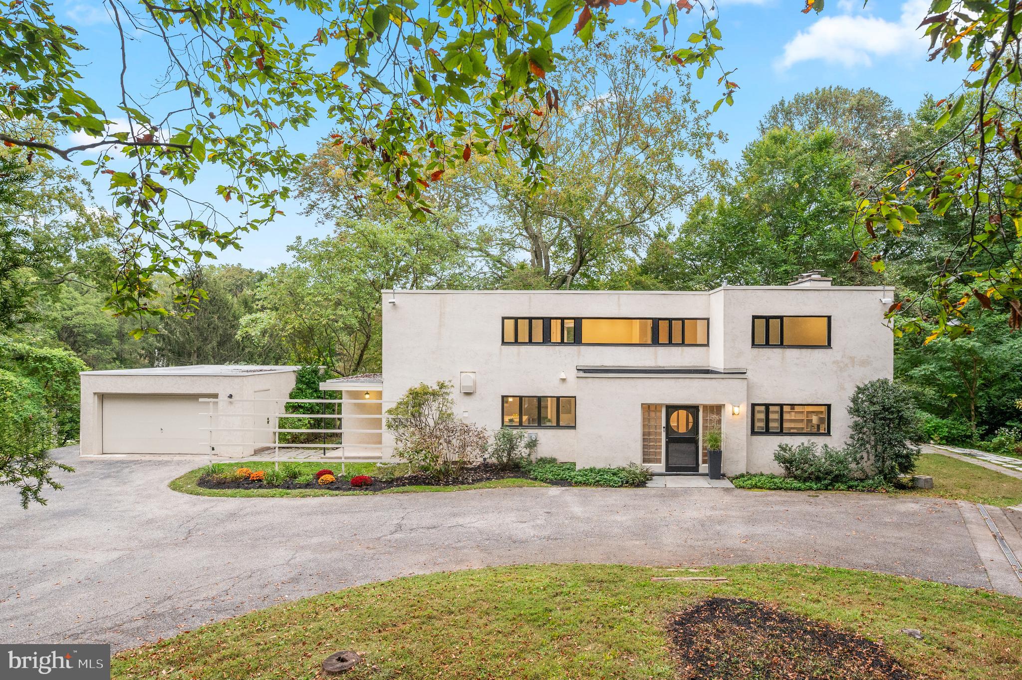 Tucked away at the end of a private cul-de-sac and set well back from Black Rock Road, this remarkable residence offers a rare combination of privacy, architectural pedigree, and natural beauty in the heart of Gladwyne. Situated on 2.6 acres within the award-winning Lower Merion School District, the home enjoys wooded views from every room and a serene connection to its surroundings. Originally designed in 1937 by architects Douglas Braik and Robert Montgomery Brown, it is a striking example of early International Style architecture, a precursor to the Mid-Century Modern movement. Defined by clean lines, flat rooflines, and expansive bands of windows, the stucco-on-masonry façade, geometric forms, and light-filled interiors embody timeless modern design decades ahead of its time.
Inside, a foyer with custom mosaic marble flooring and original metal-and-glass French doors opens to a spacious living room with wooded vistas, a wraparound terrace, floor-to-ceiling built-ins, and a fireplace. The living spaces flow into a formal dining room and a fully reimagined kitchen with custom cabinetry, quartz countertops and backsplash, Brizo fixtures, and premium appliances including a Liebherr Smart refrigerator and freezer, Fisher & Paykel Smart DishDrawer dishwasher, and Bertazzoni induction range, complemented by a light-filled breakfast nook. A home office and powder room complete the main level. Upstairs are four bedrooms and three full baths, including a primary suite with a private balcony, remodeled bath, and walk-in closet. The lower level extends the living space with a family room, laundry, and an additional full bath. A detached garage adds convenience, while renovations throughout—marble and terrazzo baths, recessed lighting, new flooring, custom cabinetry, and quartz window sills—elevate every detail of this historic home for modern living.
