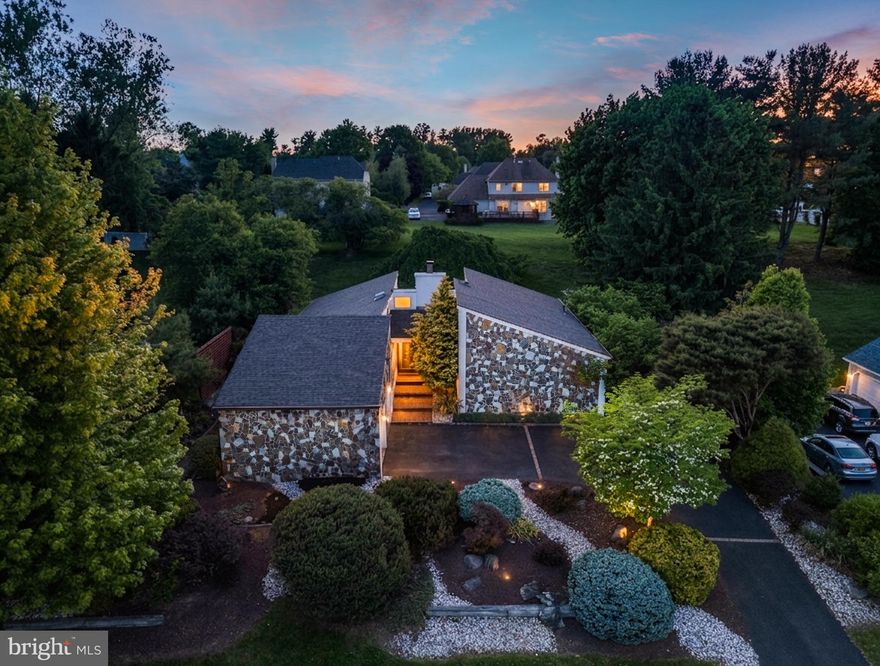 Perched atop a sloping lot and surrounded by intricate, mature landscaping, this majestic contemporary rancher offers a striking stone façade, a dramatic setting, and a truly unique floor plan. A sweeping driveway creates a motor-court feel, leading guests toward the front entry through a charming courtyard-style approach. Step inside to a welcoming foyer, where the home immediately opens into a stunning living room with soaring ceilings, expansive windows, and an abundance of natural light. Sliding glass doors extend the living space outdoors and provide direct access to the rear patio and backyard. Adjacent to the foyer, the formal dining room offers its own impressive wall of windows, along with sliding doors that lead to the side patio, creating a seamless indoor-outdoor flow ideal for entertaining. The dining room connects conveniently to the updated kitchen, which features refreshed cabinetry, a stainless steel appliance package, and excellent functionality. Also located in this wing of the home is a flexible bonus room that can serve as a fourth bedroom, private study, den, or additional family room, depending on your needs. Down a long hallway, you’ll find the private bedroom wing. Bedrooms two and three are generously sized and serviced by a beautifully remodeled hall bath featuring European-style floating vanities, large-format wall tile, and elegant brass accents. The primary suite is a true retreat, offering a grand bedroom with oversized windows, wonderful natural light, and two spacious walk-in closets complete with custom organizers and one of which features a window array and a built-in makeup table. The newly renovated private en-suite bathroom is equally impressive, featuring a European floating double vanity, large-format tile, a toilet with washlet, and a stunning frameless glass shower with brass accents and massaging wall jets. Step outside to the backyard and take in the serene setting, anchored by a majestic tree that beautifully frames the home. The expansive patio offers the perfect backdrop for outdoor gatherings, summer barbecues, and quiet evenings surrounded by nature. Do not miss this rarely offered contemporary rancher, ideally located just minutes from the shops and restaurants of Newtown and Yardley, major transit routes, and the award-winning Council Rock School District.