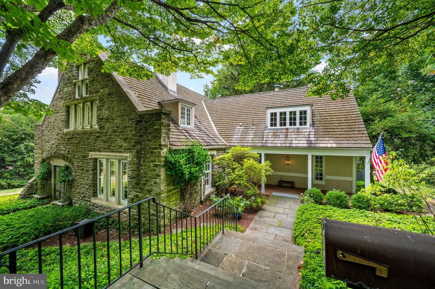 Set along one of the most picturesque and storied streets in Chestnut Hill, this extraordinary carriage house is tucked down a long private driveway, hidden beneath a canopy of mature trees and lush landscaping. A welcoming, Dutch door opens to a two-story foyer with striking black terrazzo floors with radiant heat. An arched doorway leads into the formal living room, where three oversized arched windows, once carriage entrances, fill the space with natural light. A wood-burning fireplace anchors the room, and French doors open to the backyard. To the left of the foyer, a rich, moody den envelops you in dark chocolate-painted wood paneling and beamed ceilings. A corner wood-burning fireplace with a vintage tile surround makes it the perfect place to unwind—or an inspiring home office. The great room blends comfort and function, with high ceilings, picture railing, and twin French doors to a covered side porch and patio. A long window seat runs the length of the dining nook, framed by windows. The adjacent kitchen is both beautiful and highly functional, featuring impeccable appliances: a stainless steel cooktop with six burners and griddle, Sub-Zero refrigerator and wine fridge, double wall ovens, and double GE Profile dishwashers. A reclaimed wood island offers casual seating and warmth. Beyond the kitchen, a greenhouse offers abundant sunlight and storage for gardening enthusiasts. A cozy family room at the front of the home looks into the greenhouse through a sliding glass door. A powder room and multiple coat closets complete the main level.
Upstairs, the primary suite impresses with soaring vaulted ceilings, a wall of casement windows, and an original plaster fireplace accented with hexagon terra cotta tile. The en suite bath retains its original charm and offers a spacious linen closet. A second suite sits on the opposite end of the home, also with a private bath, generous closet space, and natural light. Two additional bedrooms, a full hall bath, and a laundry room complete the upper level.
The grounds are as enchanting as the interior. Meandering brick garden paths wind through beds of pachysandra and mature perennial plantings, creating a sense of privacy and retreat. A two-tiered back patio—partially roofed, partially shaded by a pergola—provides multiple spots to dine, relax, or entertain. All of this is just steps from Wissahickon Valley Park, the shopping and dining of Germantown Avenue, and an easy drive or train ride to Center City. Steeped in history and brimming with personality, this hidden Chestnut Hill gem offers an unmatched blend of charm, privacy, and everyday convenience