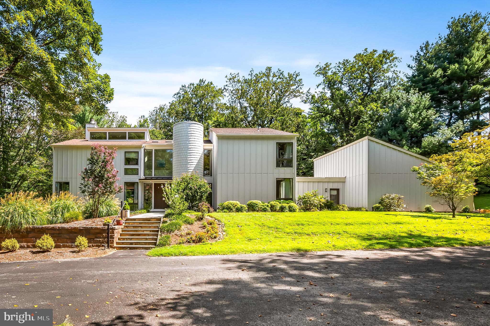 An unassuming driveway, marked only by a collection of mailboxes atop a stacked-stone planter of posies, winds up a rolling hill of perfectly scaped lawn, along a log fence shaded by a mature tree-line. Passing several of the five homes in cherished Martin’s Choice, just beyond the shared tennis court, a subtle turn leads under a canopy of fir branches and curves up to 7311 Bellona and its meticulous presentation of late 70’s mid-century modernism, giant rectangles and triangles, interconnected with symmetry, balance and the geometry of perfectly placed windows interrupting the vertical grid of its plankings’ parallel infinity. Perfect geometry continues up the flag stone steps to the pair of monolithic front doors that mirror the proportions of the plate glass above them. The 70’s were a time of open floor-plans and this foyer immediately demonstrates with sight lines to the living room, the dining room and the kitchen-adjacent den. To the left, a step down into the living room. Built-in shelves cover the rear wall, the free-standing chimney partially hides them creating a mini-library that enjoys its own sky light. A wall of floor-to-ceiling windows overlook the back deck, the opposite corners each offer the same floor-to-ceiling glass; light pours in with the trees forming backdrop of green that fill all of the frames. Back to the foyer, a 2013 addition has added a breakfast nook just beneath another wall of windows with a view of the rear patio and yard back to the wooded part of the property. A den with wood fireplace shares its space with the kitchen, a 2013 remodel with a timeless aesthetic. A laundry room (with 2nd Floor laundry chute) leads to a mud room that opens to the front walk, the back yard and the two-car garage. Off the kitchen, a separate, formal dining room has obvious potential for entertaining. The stairs, rounded by the turret, lead to the second floor’s sun-filled landing, the giant front window, the skylights, the linear intersections of railings, doorframes and rooflines continue the mathematical flawlessness. A primary suite enjoys the natural light poured from a third-level clearstory on the left.. and the four giant plate glass windows overlooking the pool on the right. A private bath, with tiled shower and soaking tub, accesses the walk-in closet. On the other side of the landing, three ample sized bedrooms line the east side of the main house. Down the hall, next to the second bedroom, is a shared full bath. Between Bedrooms Three and Four, a Jack-and-Jill joins the two. A Fifth Bedroom and Private Full Bath, created by the addition sits at the top of the stairs. Down the stairs, to the lower level, one finds a utility room to the right with storage and mechanics. To the left, past a fifth Full Bath, one finds the Walkout Family Room. With windows overlooking the side yard and the pool, this space can be a gym, a guest room, perfect for pool guests with access to the bathroom. Stepping outside, one is reminded that these grounds are more than just scenery for huge windows. A large rolling hill creates a sweeping tree-topped view from the front while the left yard feels like a shaded meadow. The right yard gets perfect exposure as it winds around the garage to the fenced back yard. Abutted by private woods, this backyard is perfect for all those backyard things: maybe a lacrosse goal or a jungle gym, a garden, or a tented party. Off the Family Room, a covered patio is ready for the grill. Off the main foyer, the cedar deck offers plenty of space for dining and entertaining. Finally, to complete the bucolic experience after a morning stroll in the rear woods, take a swim in the saltwater heated pool. With the shroud of the woods behind and acreage in front, it’s not very often that one finds a home, so well placed on a piece of land, that the comfort of seclusion comes without any of the inconvenience of distance. It feels like the middle of nowhere but in the middle of it all.