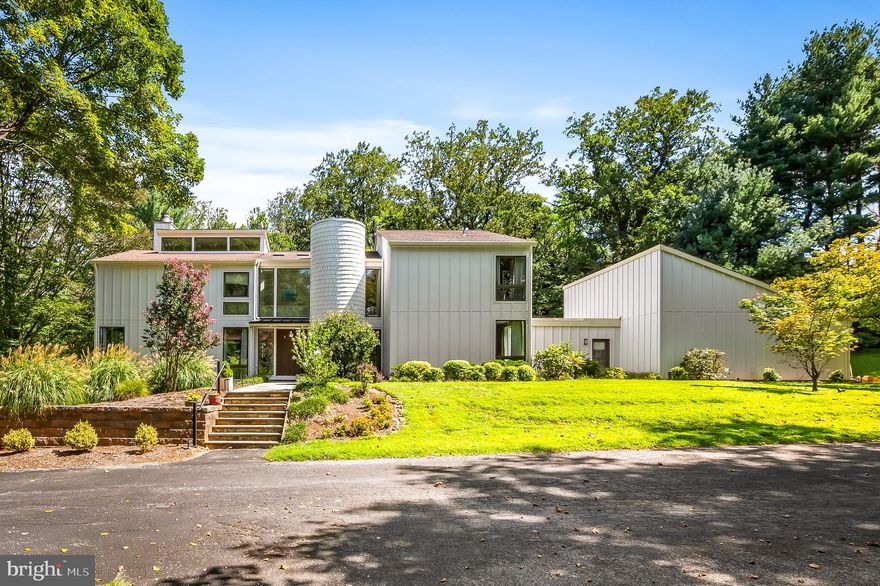 An unassuming driveway, marked only by a collection of mailboxes atop a stacked-stone planter of posies, winds up a rolling hill of perfectly scaped lawn, along a log fence shaded by a mature tree-line. Passing several of the five homes in cherished Martin’s Choice, just beyond the shared tennis court, a subtle turn leads under a canopy of fir branches and curves up to 7311 Bellona and its meticulous presentation of late 70’s mid-century modernism, giant rectangles and triangles, interconnected with symmetry, balance and the geometry of perfectly placed windows interrupting the vertical grid of its plankings’ parallel infinity.  Perfect geometry continues up the flag stone steps to the pair of monolithic front doors that mirror the proportions of the plate glass above them. The 70’s were a time of open floor-plans and this foyer immediately demonstrates with sight lines to the living room, the dining room and the kitchen-adjacent den. To the left, a step down into the living room. Built-in shelves cover the rear wall, the free-standing chimney partially hides them creating a mini-library that enjoys its own sky light. A wall of floor-to-ceiling windows overlook the back deck, the opposite corners each offer the same floor-to-ceiling glass; light pours in with the trees forming backdrop of green that fill all of the frames.  Back to the foyer, a 2013 addition has added a breakfast nook just beneath another wall of windows with a view of the rear patio and yard back to the wooded part of the property. A den with wood fireplace shares its space with the kitchen, a 2013 remodel with a timeless aesthetic. A laundry room (with 2nd Floor laundry chute) leads to a mud room that opens to the front walk, the back yard and the two-car garage. Off the kitchen, a separate, formal dining room has obvious potential for entertaining. The stairs, rounded by the turret, lead to the second floor’s sun-filled landing, the giant front window, the skylights, the linear intersections of railings, doorframes and rooflines continue the mathematical flawlessness. A primary suite enjoys the natural light poured from a third-level clearstory on the left.. and the four giant plate glass windows overlooking the pool on the right. A private bath, with tiled shower and soaking tub, accesses the walk-in closet. On the other side of the landing, three ample sized bedrooms line the east side of the main house. Down the hall, next to the second bedroom, is a shared full bath. Between Bedrooms Three and Four, a Jack-and-Jill joins the two. A Fifth Bedroom and Private Full Bath, created by the addition sits at the top of the stairs. Down the stairs, to the lower level, one finds a utility room to the right with storage and mechanics. To the left, past a fifth Full Bath, one finds the Walkout Family Room. With windows overlooking the side yard and the pool, this space can be a gym, a guest room, perfect for pool guests with access to the bathroom.  Stepping outside, one is reminded that these grounds are more than just scenery for huge windows. A large rolling hill creates a sweeping tree-topped view from the front while the left yard feels like a shaded meadow. The right yard gets perfect exposure as it winds around the garage to the fenced back yard. Abutted by private woods, this backyard is perfect for all those backyard things: maybe a lacrosse goal or a jungle gym, a garden, or a tented party. Off the Family Room, a covered patio is ready for the grill. Off the main foyer, the cedar deck offers plenty of space for dining and entertaining. Finally, to complete the bucolic experience after a morning stroll in the rear woods, take a swim in the saltwater heated pool. With the shroud of the woods behind and acreage in front, it’s not very often that one finds a home, so well placed on a piece of land, that the comfort of seclusion comes without any of the inconvenience of distance. It feels like the middle of nowhere but in the middle of it all.
