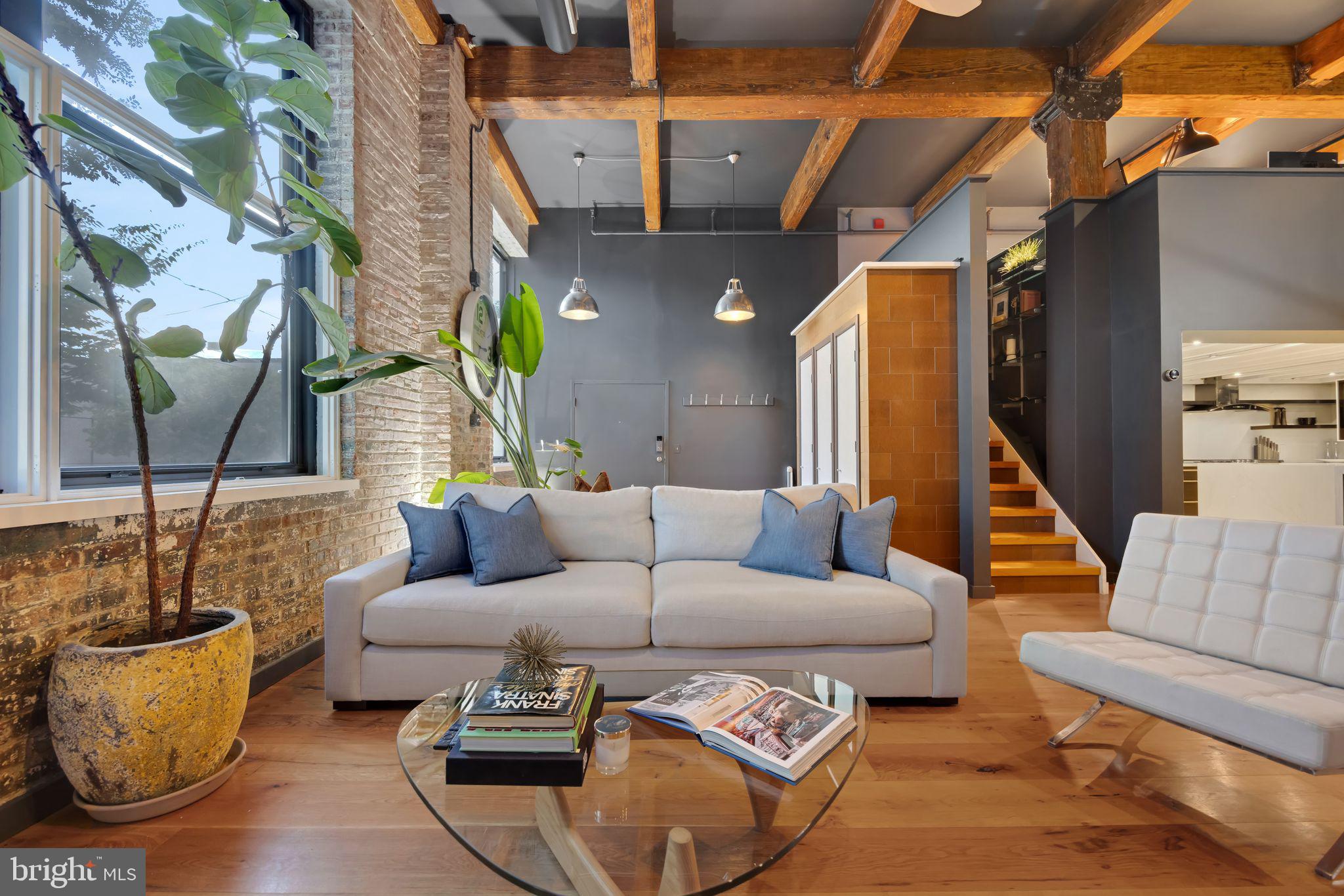 It’s not often that units like this come along! It’s even rarer to find them with garage parking (plus electric car charger) in quiet, tree-lined Bella Vista. 700 S 10th St 1A is a sleek and sexy condo located in a boutique elevator building which offers an industrial cool vibe complete with exposed brick walls, oversized wooden beams and exposed ductwork. This incredible tri-level unit has two bedrooms (one of which is lofted), upper level den/home office and two absolutely massive flex spaces. The unit door opens into a large entryway and living area with three 5 ft by 9 ft windows overlooking Palumbo Park. Notice the 9 in wide plank hardwood flooring, soaring 15-foot ceilings, custom-built coat and storage closets, and slate-colored walls that add warmth to the expansive space. There’s a sizable dining area, perfect for hosting get-togethers, beneath a modern suspended LED chandelier. There’s a crisp white open chef’s kitchen with stainless steel appliances including Samsung LED refrigerator, a brand new GE Monogram gas cooktop and GE double convection wall oven. The center island offers additional seating, a stainless steel undermount apron sink, and double thick cut Quartz veined countertops with double waterfall edges and a Bosch dishwasher. In addition to the black flat-front cabinetry, there are floating shelves flanking the modern glass range hood and a built-in shelving unit for additional storage or display. The first bedroom (which is currently set up as a nursery) is tucked back off the dining room with an oversized transom window and large closet. This private bedroom previously accommodated a queen bed set. There’s a full bath across the hall with a Carrera marble-topped floating vanity, double wide walk-in shower with subway tile and frameless glass doors and a built-in display or storage niche. The two second level rooms each have their own staircase for convenience and privacy. The primary suite is accessed through a pocket door and connects to the second full bath with two separate vanities each with abundant storage and “smart” mirrors. There’s an extra-wide walk-in shower with a rainfall showerhead, frameless glass sliding doors, and modern black Vigo fixtures. A convenient walk-in closet is located just beyond the primary bath. The other lofted room is currently set up as an office but could easily be closed off for an additional guest bedroom if needed. Last, but certainly not least, is the lower level with over 1,000 square feet to accommodate almost any living arrangement. This level is accessed via a spiral staircase off the unit entryway, or the common elevator should you need to bring in furniture, workshop equipment, large artwork or sculptures- you name it! This level also enjoys 12 foot ceilings with two extra-large rooms to configure as you please. This unit is a fantastic option for the occupant who wants living and working spaces under one roof or who desires a larger home with separate entertainment areas, and a full fitness room with the conveniences of condo-living. Last but not least, the private laundry closet/ storage closet is located across the hallway from the unit on the 1st floor and features front loading LG washer and dryer plus tons of extra storage space. Located in the heart of Bella Vista and across from an expansive park with basketball court, baseball diamond and playground, this home is located close to Whole Foods and Acme. Additionally, the Italian Market and neighborhood gems like Angelo’s, Saloon and Chapter House are just minutes away as are the Avenue of the Arts and the heart of Center City. Don’t wait to check out this one-of-a-kind home and make it your own!