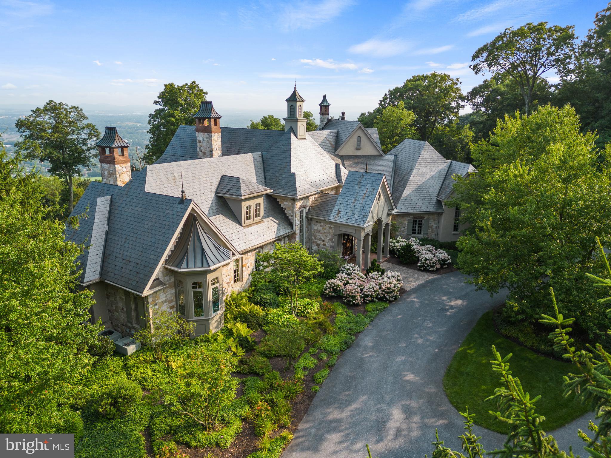 Nestled on a gorgeously manicured lot amid 10 secluded hilltop acres, this five-bedroom, 12,000+ sq-ft architectural masterpiece was lovingly designed by acclaimed architect Don Klinger to provide its owners supreme comfort and functionality while offering breathtaking views of the Susquehanna River Valley from nearly every room. With amenities too many to list (a sampling would include the five ensuite full baths plus four additional half-baths, the six fireplaces, and the 16,000-gallon outdoor koi pond with cascading waterfall), Hawkridge Estate takes mountain-side luxury living to a new level.
Situated at the end of a private, wooded drive within a gated community, the mansion features an all-natural stone façade and grey slate roof, with a spacious circular driveway and a broad double garage cleverly hidden to the side. The lofty entrance portico guides visitors into a stately hand-chiseled limestone foyer adorned with shimmering chandeliers and a sweeping, wooden grand staircase that curls upward to the second-floor bedrooms and a luxurious executive office suite.
Warm, sophisticated opulence characterizes every room of this three-story home, from the Great Room’s massive 24’-high beams and 1800-lb, no-seam rug imported from Thailand, to the elaborately hand carved millwork of the formal dining room. The expansive chef-quality kitchen features 200-year-old white oak floors, two-tiered solid granite counters, more than ample cabinet space, and a walk-in pantry. The regal owner’s retreat includes oversized closets and a jacuzzi surrounded by stained-glass windows. Hand-painted decorative tray ceilings throughout the house highlight natural motifs of plants, florals, and wildlife, elegantly complementing the house’s soothing earth-tone palette and Country French interior theme. The result is a retreat that is both luxuriously comfortable and hospitality ready.