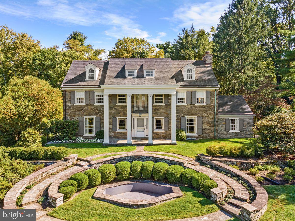 This stately Georgian Revival house, with true Wissahickon Schist stone throughout, along with its lovely grounds, is the anchor of its coveted Morris Arboretum neighborhood, and offers the utmost privacy.
This property extends the beauty of the neighboring Morris Arboretum with mature trees and a garden that has been lovingly maintained by its owner. The house is equally serene. Enter up curved slate pathways surrounding a front fountain to the two-story portico with columns and a Juliet balcony. The gracious front hall has beautiful, wide planked wood floors that continue in the living and dining rooms. The spacious living room to the right has a stone fireplace and regular and floor-to-ceiling windows allowing for maximum light. Next to it, is a lovely study/den that was added in the 1980s also with floor to ceiling windows and a sliding glass door that opens to the magical backyard. To the left of the front door is an equally gracious dining room, with windows on three sides. Next to the dining room is the morning sun-drenched kitchen with a door leading to the back stone patio expansion built in 2000. From the Patio, take in the splendid
in-ground pool with gorgeous perennial flowers that is the mainstay of the pristine and lovely yard that also hosts a grilling area above right of the pool in its own terraced space.
Back in the house and upstairs on the 2nd floor you will find three spacious bedrooms and two bathrooms. Two of the bedrooms share a Jack and Jill bathroom and at the top of the stairs is a wonderful landing that looks out over the neighborhood. Three more bedrooms and two baths await you on the 3rd floor. This well-loved and maintained home has a large two car garage with access to the cellar, oil heat, a buderus heater, radiators and storm water drains,
This home, offering 5 plus bedrooms and 4.2 baths, is a true Chestnut Hill Gem. It is wonderfully elegant inside and out. It is also minutes away from all that Germantown Avenue offers with its great shopping and dining, Septa trains, schools and the trails of the Wissahickon Valley, not to mention the Morris Arboretum. This house is not to be missed!