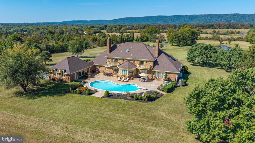 Truly extraordinary and rarely available. Set on 19.9 breathtaking acres in the Log Mill Road area of Prince William County, this all-brick Tudor-style estate is a rare jewel surrounded by open fields and framed by sweeping views of Bull Run Mountain. To the front, a stocked pond and gently sloping meadows enhance the setting, while to the rear, a Caribbean-blue pool and expansive outdoor entertaining area create a private enclave perfect for family gatherings, pool parties, and Sunday brunches by the pool. A tree-lined driveway leads to a circular approach and introduces the home with presence and elegance. This property is exceptional not only for its location and scale, but also because it has no HOA and no conservation easements, providing total freedom for your lifestyle and future plans.

Inside, a grand two-story foyer welcomes guests with marble floors, a sweeping circular staircase, and a gracious keeping/overflow area perfect for greeting visitors or allowing guests to linger at the end of an evening. The main floor showcases intricate moldings, including dental and ornate details, highlighting the craftsmanship throughout.

The kitchen is a chef’s dream, featuring granite counters, custom cabinetry, and spectacular views of the pool. The formal dining room with fireplace offers a warm and inviting space for entertaining. The family room, positioned close to the kitchen, features its own fireplace, access to a conveniently located bathroom, and walks out to the pool while leading seamlessly into the entertainment room.

The entertainment room is a true showstopper: huge in scale with marble floors, direct pool access, a full bar, and a full sports bar that overlooks the TV—perfect for watching the Masters, the Super Bowl, or your favorite Netflix series. This space is designed for hours of joy, whether hosting Sunday night movies, game-day celebrations, or large-scale gatherings. Its scale feels grand, yet the layout allows for a warm, inviting atmosphere where friends and family can relax and enjoy every moment.

On the opposite side of the home, a full-sized library or executive office awaits, complete with built-in bookshelves, a full fireplace, and access to the pool—an inspiring space for work, reading, or quiet reflection. Adjacent is a guest suite that is both enormous and cozy, offering a private retreat with ensuite bath and stunning pond views.

Upstairs, the primary suite is a true retreat, featuring a private balcony with views of the pool, windows that capture dramatic sunset views over the mountains, his and her bathrooms, enormous his and her walk-in closets, a fireplace, and a generous sitting/dressing room. Two additional bedrooms on the upper level are equally impressive, each with ensuite baths and oversized walk-in closets. One bedroom also has a balcony, offering views of the pool. The princess balcony overlooking the foyer is equally stunning, with intricate moldings continuing all the way up the two-story space, enhancing the dramatic architectural presence of the home.

The lower level provides even more lifestyle space with a recreation room, finished storage, a gym with sauna, and a large laundry/workshop area complete with a laundry chute. Completing the home is an impressive 3-car side-load garage, providing both function and curb appeal. The home is built with incredible solid construction that inspires confidence while still feeling welcoming and livable.

With its combination of wide-open acreage, pond, pool, mountain vistas, and extraordinary interior spaces, this estate is truly an impossible find. Located just 3 minutes from Loudoun County and close to local breweries, the town of Middleburg, and all the amenities of Haymarket. The lot is the perfect canvas—with room for a barn, an indoor riding arena, a driving range, or an enormous garden.  For those who love to fish, the stocked pond is ready to provide  hours of enjoyment.  All offers due by Tuesday, October 7 at  2:00 PM