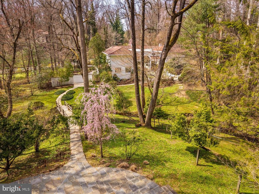 Few properties in Lower Merion combine this: 1.62 private acres, more than 8,000 square feet, and six bedrooms within minutes of Center City. This six-bedroom, 5.5-bath residence was designed for large-scale entertaining. The main level opens through a dramatic gallery entry - brick walls, exposed beam ceiling, marble floors - into a great room of nearly 36 by 21 feet, finished in polished marble with a crystal chandelier and French doors to the rear gardens and fountain patio. A step-down family room centers around a hand-carved walnut bar with iron scrollwork and marble top - a true architectural centerpiece. The 22x22' kitchen features granite countertops, dark wood cabinetry, and a hand-hammered copper hood set against a full-height brick wall. A breakfast room, catering room with bar and wine storage, private office with cherry built-ins, and two-stop residential elevator complete the main level. Six large bedrooms and four full baths upstairs, including a primary suite with marble bath and dressing room. Finished lower level. Auxiliary generator Swimming Pool. Fountain patio.