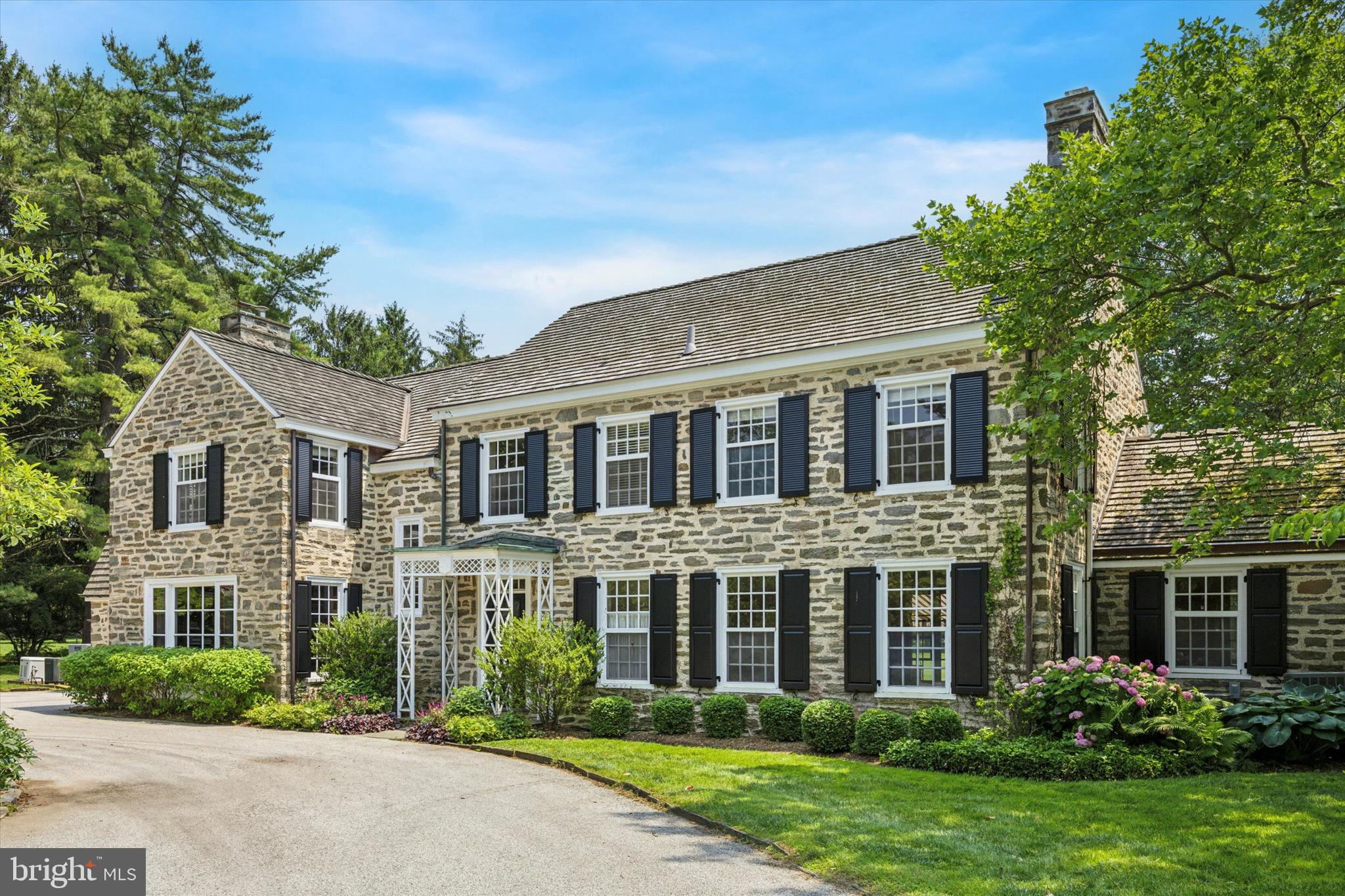 Tucked at the end of a long, winding private drive, 1024 Spring Mill Road in Villanova, PA, unveils a stately stone colonial, a haven of privacy and understated comfort. on lushly landscaped grounds. Surrounded by preserved land, this 6-bedroom, 5-full-and-1-half-bathroom retreat is enveloped by vibrant flowerbeds, mature trees, and native shrubs, creating a secluded oasis. For those who cherish solitude and space, this home is the desired timeless sanctuary.
The traditional floor plan harmonizes connection and seclusion, with floor-to-ceiling glass windows and French doors flooding each room with natural light and framing tranquil views of the grounds. The living room, warmed by a wood-burning fireplace, invites cozy gatherings and opens to a large terrace covered by an awning, extending off both the living and dining room. This shaded terrace, ideal for outdoor dining and relaxing out of the sun, enhances the home’s indoor-outdoor flow. The dining room offers ample space for intimate meals or family celebrations. A private den, with its own wood-burning fireplace, provides a quiet retreat for work or reflection. The great room, featuring another wood-burning fireplace and flowing directly from the large chef’s kitchen, serves as the heart of daily living. The kitchen, a functional centerpiece, boasts a 6-burner Dacor stove, two Bosch ovens, a Bosch dishwasher, a Sub-Zero side-by-side refrigerator/freezer, and an expansive marble countertop with seating for four, perfect for casual dining. A mud room hall leads to a practical office and laundry area, ensuring effortless organization. The pool room, with a bar, full private bath, and its own wood-burning fireplace, offers a versatile space for leisure or hosting by the very private pool.
Upstairs, the primary suite is a private sanctuary, featuring his-and-her bathrooms, an extensive walk-in closet, and serene views of the tree-lined grounds, ensuring restful seclusion. Four additional bedrooms, served by two well-appointed full bathrooms, provide generous space for family, guests, or home offices, each offering internal privacy for quiet moments. The thoughtful layout ensures everyone has their own retreat, from the den’s solitude to a bedroom’s cozy nook.