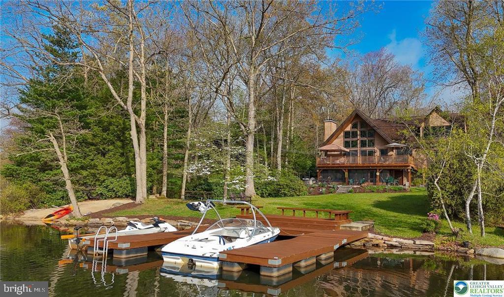Imagine celebrating each glorious Pennsylvania season from a home that defines idyllic lake life, comprehensively enjoyed from its private beach or sprawling paver patio, the twenty-foot-tall living room windows or the private upper-level sanctuary of the primary bedroom. Approaching from the home’s front, the attention to detail and care are immediately evident in the warm tones and stone accents wisely chosen to harmonize in the lush sylvan surroundings, protected greenbelt area borders the property to one side. The entry porch opens to a lovely foyer, its ceramic tile becoming gleaming hardwoods leading up to the main level’s free-flowing gathering space. The kitchen is open to a gently defined dining room as well as the living room, its wood-beamed, pine tongue and groove cathedral ceilings soaring to a twenty-two-foot peak. Abundant windows overlook the deck, yard, and lake beyond while a wood-burning fireplace offers charm and comfort. A stone fireplace surround with wood accents in trim and railings offer organic touches and, when paired with the floor and ceilings, appropriately hearken the perfect feel of a cozy lakeside cabin. Secluded from the gathering spaces are two well-sized bedrooms, one with a built-in home office workstation and beamed ceiling, the other a beamed, tongue and groove ceiling amplifying its rustic beauty. A main-level full bath is fully updated with a lovely tub-size shower. The main level also boasts a welcome number of closets in the hall and bedrooms. A staircase leads from living room to the upper level where a generous sitting room loft awaits, overlooking the living room, it is the perfect nook from which to enjoy the soaring windows with their views of the glistening lake. A second, updated full bath accompanies two lovely bedrooms, both taking full advantage of their dormer location to continue the wood ceilings to offer an ideal sylvan sleeping experience. The primary bedroom offers four hundred square feet with the gem in this crown being a large window situated in the dormer allowing for perfect views of the lake and wooded hills to the south; imagine waking to this view every morning and curling up in a comfy chair with your coffee as the first beams of sun glitter on the gentle rippling of Lake Hauto. The primary bedroom also offers two closets, one a generous 9x14 walk-in, as well as a 150sf private home office. The home’s lower level offers abundant entertaining space from its five-hundred square foot L-shaped family room set around a two-hundred square foot second kitchen with laundry facility. An enclosed porch offers access to the yard and paired with the lower level’s utility room, storage space for a multitude of outdoor toys and furniture! Access to the home’s backyard amenities is also found in the main-level composite deck. Sliding glass doors from the living room offer access to this area with ample space for an outdoor living room and outdoor dining room! Stairs lead down to the 858 square foot paver patio which offers a bonus in its regulation, concrete shuffleboard court as well as a corner nook with fire pit, a stone wall offering an attractive border with seating potential. The patio gives way to a lawn gently sloping to the home’s private beach to the right, and three-boat-dock to the left with a lovely gathering space, built in benches an ideal location to enjoy the water’s edge. The lake front of this home is the southernmost point of land extending into Lake Hauto, offering an ideal location to take in the complete scope of its riparian beauty. While Lake Hauto Club does allow for powerboating, jet skis, and similar, this lake side oasis sits within the “no-wake zone” of the lake, offering a more tranquil experience. Lake Hauto Club amenities include four public beaches with boat docks and pavilions, tennis and pickleball courts, a community gym and library, and of course the 330-acre lake itself. Situated at the mid-point bet