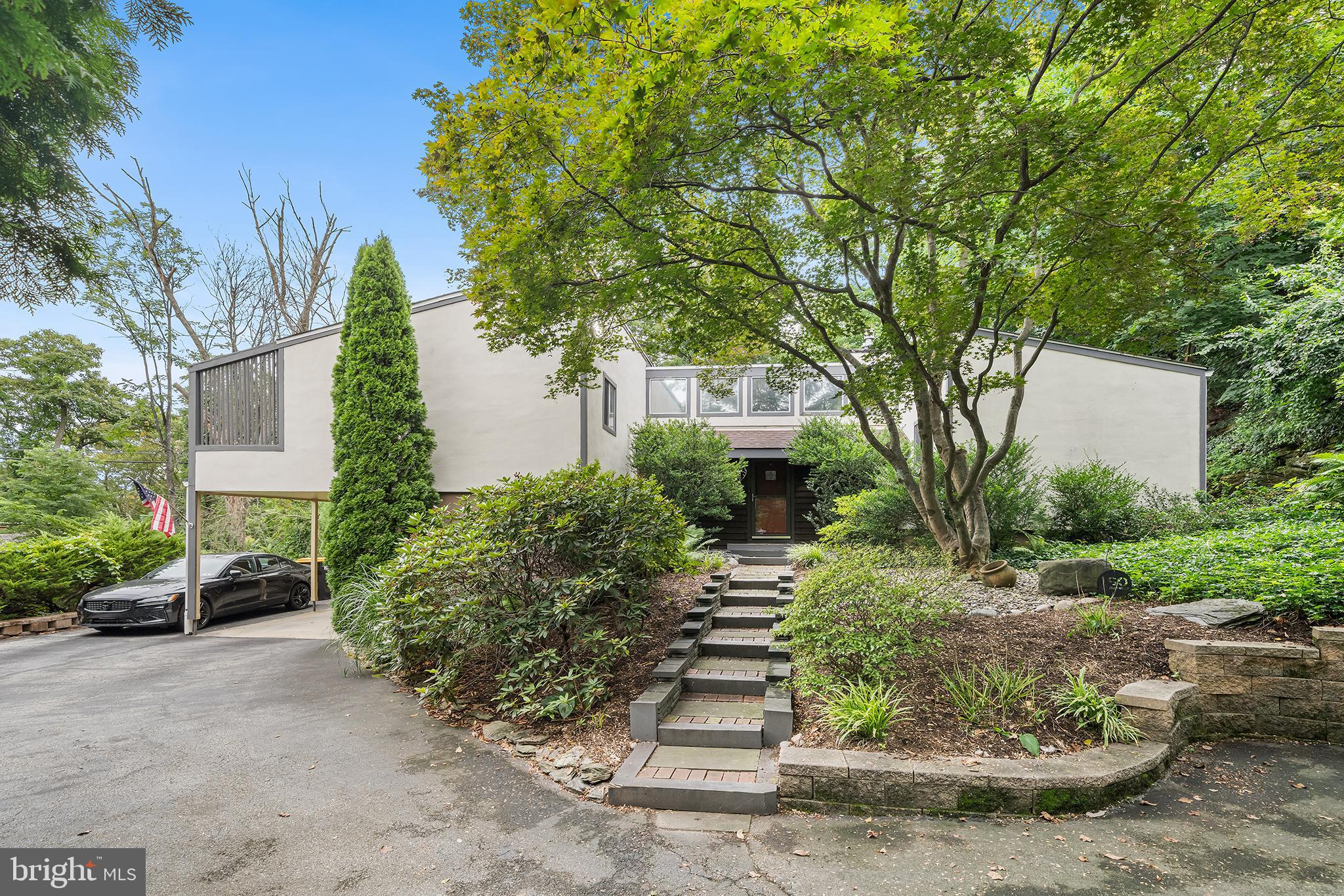 Situated at the end of a quiet cul-de-sac and enclosed by mature trees and landscaping, this contemporary home offers a wonderful sense of privacy and a layout that is open, inviting, and apt for entertaining. The first impression is from the private driveway, where the home’s modernist design is on full display. Bold geometric form, angular roof sections, and contrasting facade materials distinguish the property. A landscaped stone pathway leads to the covered front entryway, which is set beneath a statement wall of windows. Inside, the custom design and soaring, beamed ceilings draw you into an open entertaining area. A few steps down from the entryway landing, a sunken living room is framed by a dramatic floor-to-ceiling stone fireplace—an inviting space that naturally flows to all other areas of the property. The dining room is adjacent to the living room, between the kitchen and an oversized deck with sliding glass doors. The property’s connection to the outdoors can not be overstated; there is incredible continuity between the living areas and the back deck, making hosting a breeze. The windowed kitchen is complete with granite countertops, stainless steel appliances, and a convenient breakfast bar. The residential quarters are a few steps up or down from the open entertaining space. The upper level contains three spacious bedrooms and two full bathrooms. Chief among them, the primary bedroom is positioned at the back of the house, and offers the owner a private deck behind sliding glass doors, a walk-in closet, and an en-suite windowed bathroom with a glass-framed shower. The primary suite comes complete with a small bonus room, which can suit a number of uses! Two additional bedrooms and a full hallway bathroom with modern tiling and a tub/shower enclosure complete the property’s upper level. The lower floor is well utilized, with an additional bedroom (for 4 in total) and a half bathroom. This floor also offers a bonus flex room, laundry room with storage cabinets and a wash basin, and a door leading directly to the carport and side yard! Before heading out, spend some time on the tiered back deck, which runs the length of the dining room and living room and connects the back of the house to the side yard. Situated within the Upper Merion Area School District, the home scores big on convenience—it’s minutes from the King of Prussia Mall, top-notch dining, major highways, and the SEPTA Gulph Mills station. The proximity to Center City, the Philadelphia International Airport, and local attractions makes this property as connected as it is peaceful. Disclaimer: Compass RE and Evan Frisina do not guarantee square footage estimates or real estate tax data. The square footage estimate is inclusive of the lower level.