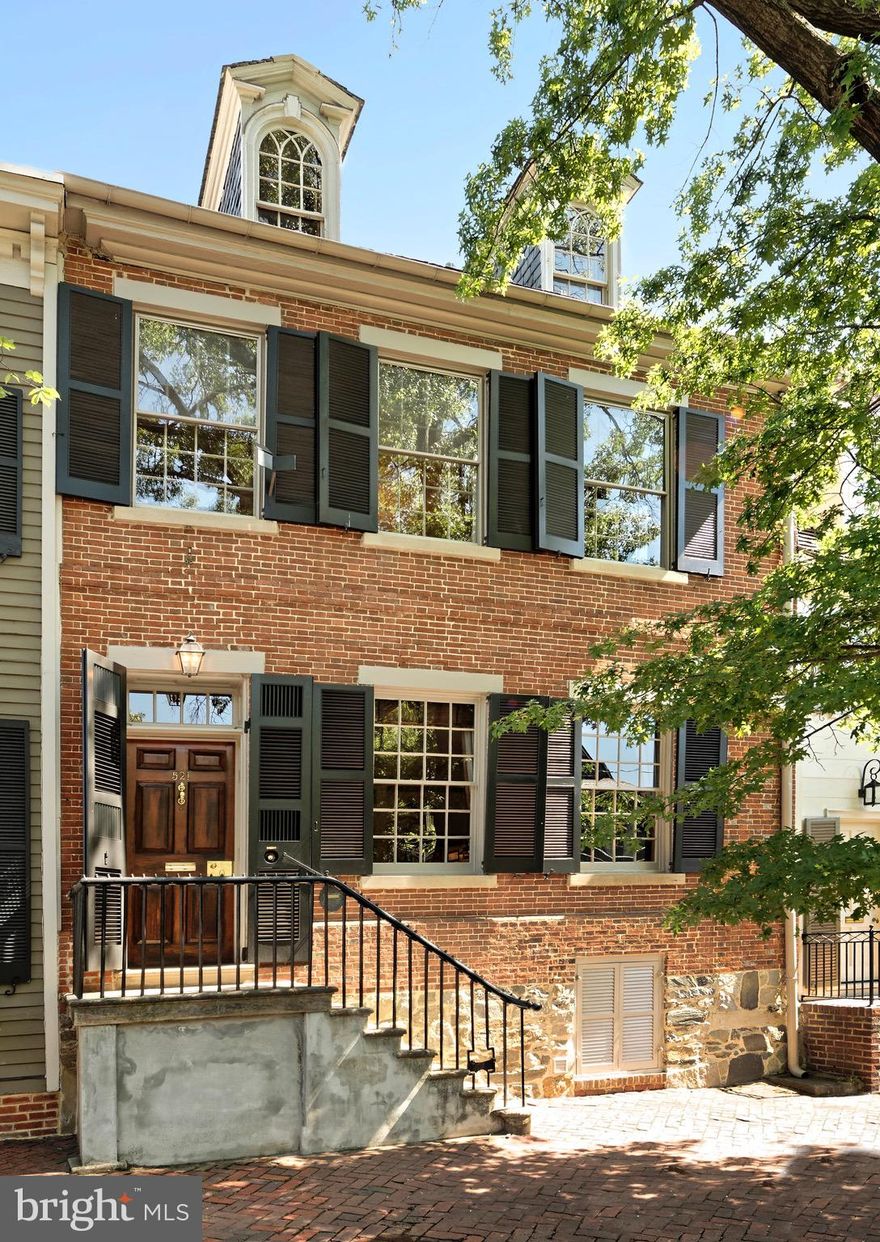 First time on the market in 35 years, this important, historic house will amaze you.  Built in 1789 by Lawrence Hooff, Sr., this distinguished brick townhouse was once home to George Washington’s nephew and heir to the Mount Vernon Estate, Bushrod Washington, who served as Associate Justice of the Supreme Court and presumably used the two front rooms as his law offices and the upper levels as the family living quarters.  Today, this fine 4 bedroom, 3.5 bath residence embodies the historic beauty of yesteryear with modern conveniences including generously proportioned rooms, original hardwood floors, 12 over 12 windows, exquisite built-ins, off-street parking, and a wonderful private patio.
This timeless home greets you with an airy entrance hall boasting 10’ ceilings graced by an elegant hand carved archway. Entertaining is effortless in the spacious library featuring a wood burning fireplace, oversized windows, and custom bookshelves.  A stately dining room with French doors overlooking the courtyard is a lovely setting for gatherings. It is believed that President George Washington dined here with his nephew.  And in recent years President Bill Clinton dined here.  Prepare meals in the well-equipped kitchen enriched by a dramatic exposed brick wall and beamed ceiling.  Set conveniently off the kitchen is a handsome family room featuring paneling from a barn in southern Virginia built in 1800, a fireplace and a glass door opens to the outdoors. A powder room is off the front hall to accommodate guests.  
Above the library sits a second-floor parlor showcasing the unparalleled craftsmanship of long ago.  It is   a vision of beauty with its arched entrance doorway and original glass fanlight, tall windows, fireplace, and built-in cabinetry setting the perfect tone for formal entertaining.  A charming primary bedroom featuring original paneling and millwork, fireplace, and heart of pine floors provides an elegant respite. Additional second floor highlights include a light-filled guest suite, updated marble bath, and a large cedar closet. The third level includes 2 sunny bedrooms, a full bath and desk space.
Surrounded by old brick walls, the delightful patio is adorned with a mature magnolia tree, roses, holly, and an espaliered pyracantha offering the perfect setting for al fresco dining. One off-street parking space for this property  is located beyond the gate.