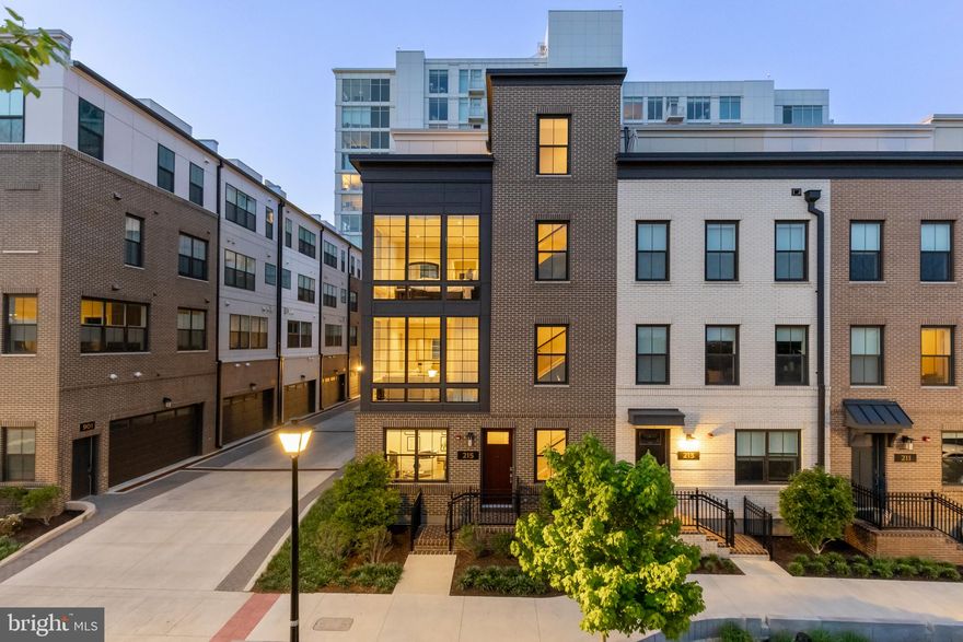 Gracing a premium corner lot in Alexandria's sought-after Venue community, 215 Montgomery is a timeless four-level residence just steps from  Potomac River and moments from the  charm of Old Town—The Venue is a masterfully planned enclave of luxury townhomes and condominiums developed by CARR Companies. Positioned on a premium corner lot, 215 Montgomery Street is a stately four-level residence clad in brown brick on all sides, evoking a contemporary reinterpretation of a classic Upper West Side brownstone.

At 24 feet wide and offering 3,344 square feet of elevated living space, the home boasts one of the largest layouts in the community—evoking the sensibility of single-family living within a walkable urban core. A gracious foyer introduces the home, with a private den ideal for welcoming guests, a formal powder room, and access to the extra-wide, attached two-car garage—equipped with EV charging and custom storage solutions.

Ascend via the sweeping staircase or all-level private elevator to the main level, where soaring ceilings and two walls of glass bathe the open-concept floor plan in natural light. Wide-plank oak hardwoods unify the expansive living area, anchored by a striking double-sided fireplace that subtly delineates formal and casual spaces. A second powder room adds additional convenience.

At the heart is a chef’s kitchen designed with clean lines and premium finishes—white shaker cabinetry, a River Rock center island, Calacatta Neve quartz countertops with matching slab backsplash, and a fully integrated Bosch premium appliance suite. A French door wine cooler adds a refined touch.

The upper-level primary suite is a true sanctuary, spanning half the floor and featuring dual custom walk-in closets and a dedicated dressing area. The lavish bath showcases a double River Rock vanity, Bianca Carrara matte flooring, a large glass-enclosed shower, and a separate water closet. Past the side-by-side laundry closet, a secondary bedroom enjoys its own ensuite bath with chic tiling and a walk-in glass shower.
The top level offers a third bedroom with ensuite bath, a cozy loft lounge, and access to the home’s pièce de résistance—a private rooftop terrace with a custom kitchen and  panoramic views of the Potomac and the National Harbor, MD skyline.

Smart home features abound, including Ubiquiti Networking and Savant Home Automation, Ring security, Sonos in-ceiling audio, Lutron motorized window treatments, Ecobee smart thermostats, Samsung, Sony and LG wall-mounted TV's, and Kohler electronic bidets throughout.

Set moments from the Mount Vernon Trail, MetroStage, and Old Town’s beloved shops and eateries, 215 Montgomery Street presents an unrepeatable opportunity to enjoy an innovative, luxurious urban lifestyle in the heart of Alexandria.