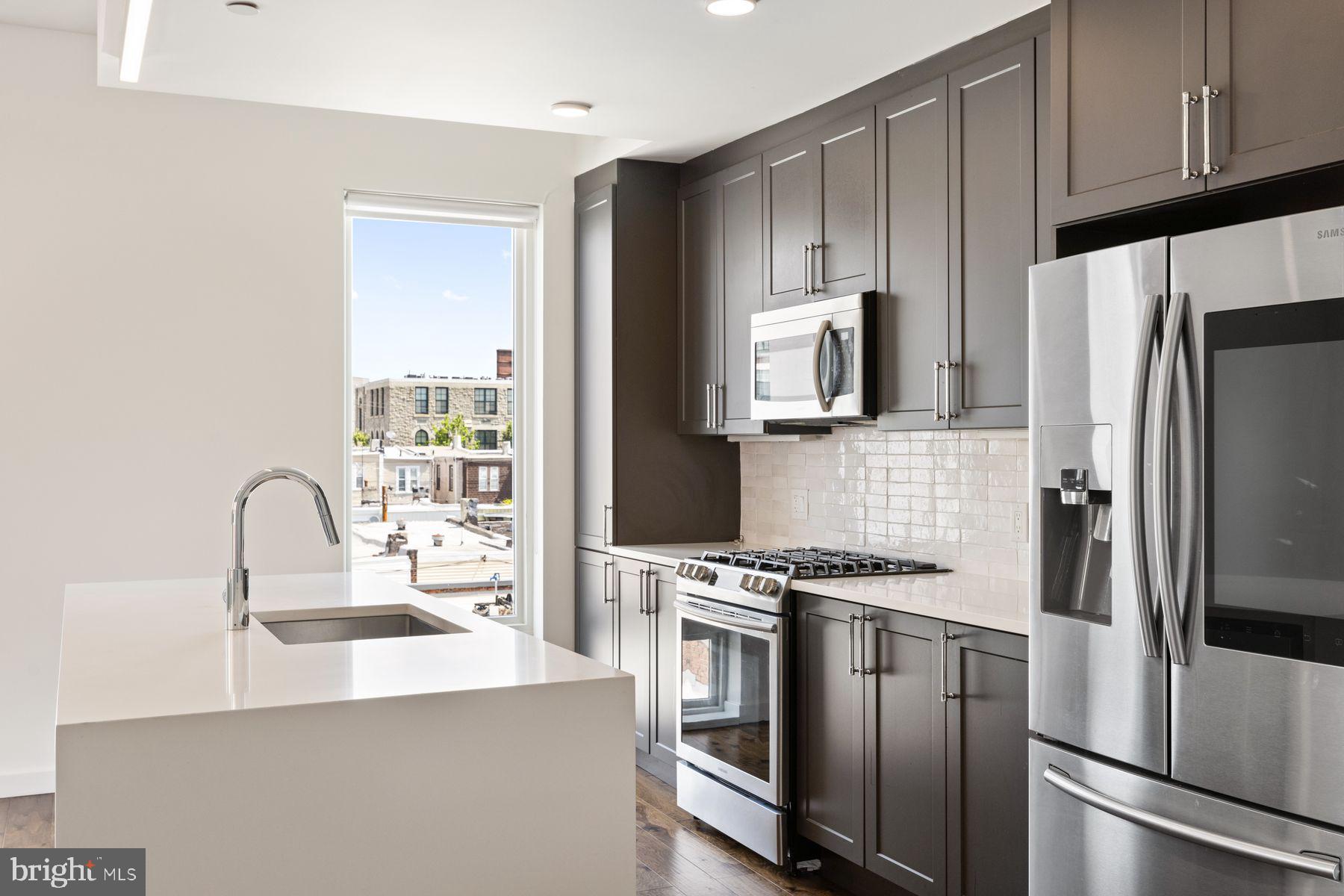 Welcome to Penthouse PH4 at Otto On The Park, a top-floor 2-bedroom, 2-bathroom corner unit offering 1,256 square feet of refined living space and a sprawling 1,300-square-foot private roof deck with panoramic Center City views. Sunlight pours through 7-foot-tall windows, illuminating the open-concept layout that seamlessly connects the smart kitchen—featuring a waterfall island and Samsung Family Hub refrigerator—to the main balcony, perfect for entertaining. The primary suite boasts a walk-in closet and a luxurious en-suite bathroom with dual vanities, a glass-enclosed shower with dual sprayers, and a stone bench. The second bedroom also includes a walk-in closet and is served by a tiled hall bath with a soaking tub. Additional amenities include a laundry closet, coat closets, and an open office nook. Residents enjoy keyless building access, a Nest thermostat, and one deeded, gated parking space. Situated in Brewerytown, PH4 is within walking distance of local favorites like Rival Bros Coffee, Baby's Kusina + Market, New June Bakery, and Harper's Ice Cream, offering a neighborhood experience unlike most!