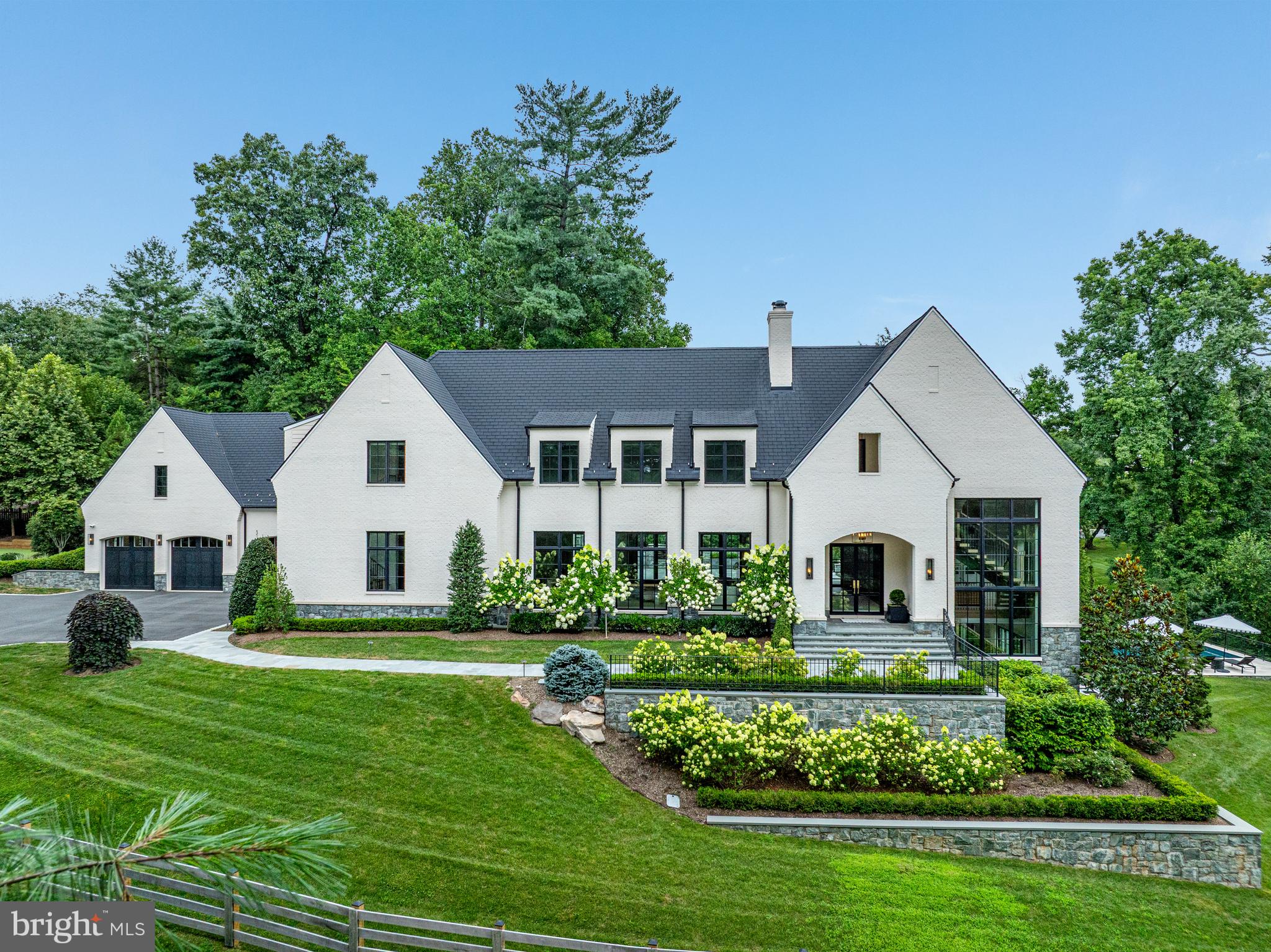 This sophisticated Modern Estate blends timeless European-inspired architecture with contemporary elegance, set on a beautifully manicured private lot in the exclusive cul-de-sac enclave of The Lane on Georgetown Pike adjacent to McLean’s historic Langley Farms.
Striking in both form and proportion, the home commands attention with its clean lines, harmonious symmetry, and soft pastel brick façade, accented by oversized black casement windows and a dramatic steel entry door. Locally quarried Carderock stone wraps the foundation, grounding the residence in Virginia’s rich architectural tradition, while layered stone retaining walls and curated landscaping lend a stately presence to the grounds.
A gracious Foyer introduces the home’s refined interiors, anchored by a sculptural floating staircase bathed in natural light from a soaring window and illuminated by unlacquered brass pendant lights. Throughout the main living spaces, rift and quarter-sawn white oak floors convey warmth and craftsmanship, complemented by an Elevator servicing all three levels for seamless access.
The Living Room centers around a bold marble fireplace, while the Dining Room, framed by custom sliding doors, opens to a fully appointed Butler’s Pantry. The minimalist Kitchen is a masterclass in design featuring top-tier appliances, custom cabinetry, and dual center islands flowing effortlessly into a light-filled Family Room with a sleek modern fireplace as its focal point.
Upstairs, the Owner’s Suite is a serene sanctuary with soaring cathedral ceilings and two generous dressing rooms, one outfitted with a bespoke vanity. The spa-like marble Bath exudes luxury, with dual oversized vanities adorned by dramatic waterfall countertops and Waterworks brass fixtures. A beautifully lit Gallery leads to three additional ensuite Bedrooms and a Laundry Room, each space echoing the home’s refined aesthetic.
The sun-drenched Lower Level is equally impressive an expansive space designed for modern living and entertaining. A double-sided fireplace open to the Recreation Room and Game Room, while a sleek Wet Bar and glass-enclosed, temperature-controlled Wine Cellar provide the perfect backdrop for gatherings. Additional highlights include an Exercise Room, private Bedroom Suite, a versatile Bedroom/Massage Room with access to a tranquil Spa Bath and steam shower.
From the Lower Level, French doors open to a serene Stone Terrace with a built-in Firepit and Pool ideal for lounging or evening entertaining. A stone staircase ascends to the dramatic Upper Terrace, which features an Outdoor Kitchen, Stone Fireplace, and sculptural Fountain creating a seamless indoor-outdoor living experience that is both elegant and inviting. To complete the outdoor living area there is gracious green space with outdoor seating and a separate putting green fun for family and friends.