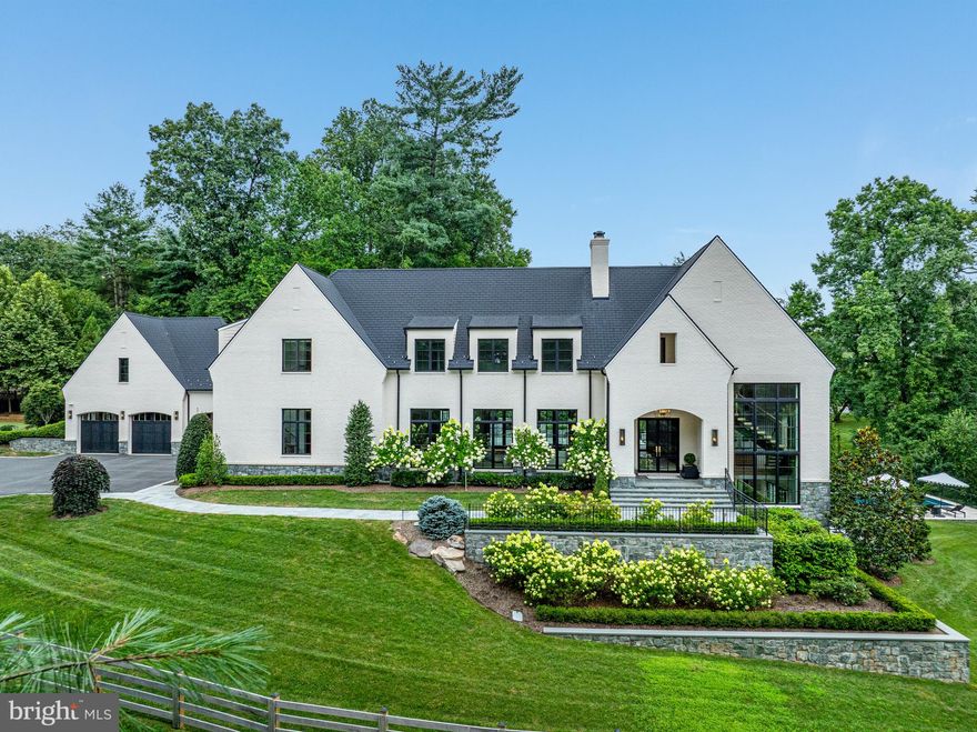 This sophisticated Modern Estate blends timeless European-inspired architecture with contemporary elegance, set on a beautifully manicured private lot in the exclusive cul-de-sac enclave of The Lane on Georgetown Pike adjacent to McLean’s historic Langley Farms.

Striking in both form and proportion, the home commands attention with its clean lines, harmonious symmetry, and soft pastel brick façade, accented by oversized black casement windows and a dramatic steel entry door. Locally quarried Carderock stone wraps the foundation, grounding the residence in Virginia’s rich architectural tradition, while layered stone retaining walls and curated landscaping lend a stately presence to the grounds.

A gracious Foyer introduces the home’s refined interiors, anchored by a sculptural floating staircase bathed in natural light from a soaring window and illuminated by unlacquered brass pendant lights. Throughout the main living spaces, rift and quarter-sawn white oak floors convey warmth and craftsmanship, complemented by an Elevator servicing all three levels for seamless access.

The Living Room centers around a bold marble fireplace, while the Dining Room, framed by custom sliding doors, opens to a fully appointed Butler’s Pantry. The minimalist Kitchen is a masterclass in design featuring top-tier appliances, custom cabinetry, and dual center islands flowing effortlessly into a light-filled Family Room with a sleek modern fireplace as its focal point.

Upstairs, the Owner’s Suite is a serene sanctuary with soaring cathedral ceilings and two generous dressing rooms, one outfitted with a bespoke vanity. The spa-like marble Bath exudes luxury, with dual oversized vanities adorned by dramatic waterfall countertops and Waterworks brass fixtures. A beautifully lit Gallery leads to three additional ensuite Bedrooms and a Laundry Room, each space echoing the home’s refined aesthetic.

The sun-drenched Lower Level is equally impressive an expansive space designed for modern living and entertaining. A double-sided fireplace open to the Recreation Room and Game Room, while a sleek Wet Bar and glass-enclosed, temperature-controlled Wine Cellar provide the perfect backdrop for gatherings. Additional highlights include an Exercise Room, private Bedroom Suite, a versatile Bedroom/Massage Room with access to a tranquil Spa Bath and steam shower.

From the Lower Level, French doors open to a serene Stone Terrace with a built-in Firepit and Pool ideal for lounging or evening entertaining. A stone staircase ascends to the dramatic Upper Terrace, which features an Outdoor Kitchen, Stone Fireplace, and sculptural Fountain creating a seamless indoor-outdoor living experience that is both elegant and inviting. To complete the outdoor living area there is gracious green space with outdoor seating and a separate putting green fun for family and friends.