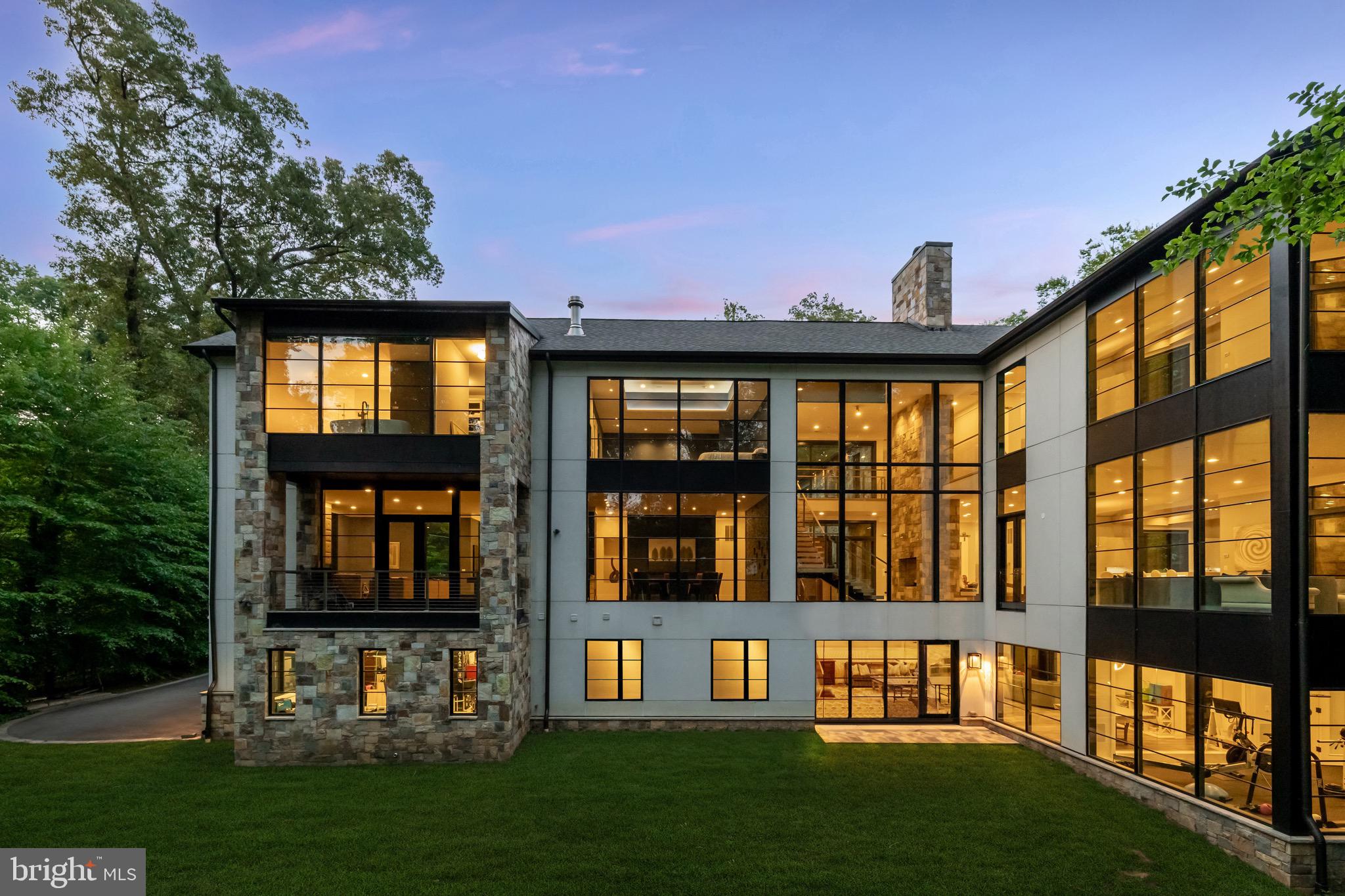 Tucked away, just off the convenience of Massachusetts Avenue, lies idyllic Bay Tree Lane, adorned by lovely homes on large lots and under the canopy of gorgeous mature trees. Perfectly positioned on the best lot of the quiet cul-de-sac, is the stunning contemporary estate of 3 Bay Tree Lane. Constructed in 2018, this 12,700 square foot masterpiece stands at the epitome of cutting-edge design and masterful craftsmanship. The distinctive facade is clad in an effortless combination of natural stone, mahogany, stucco, and lit by swirling gas lanterns, a combination that make this home feel like it could just as easily be mountainside at an exclusive ski resort.
Arranged in an L-shape, the home is approached by two separate driveways leading to an upper three-car and lower two-car garage for the automobile enthusiast. The arrival parking pad is generous in scale and provides a grand, flagstone approach to the main entrance. An oversized mahogany door pivots open to reveal the grand foyer. Effectively a glass section bridging two separate masses, the feeling is as if you are standing in the surrounding nature of the home. The double-height entryway is flooded with natural light on both sides via floor to ceiling windows with motorized blinds, illuminating the 8-inch quarter-sawn white oak floors. A functional work of art, the floating staircase is framed with tempered glass and stainless steel.
Opposite is a towering two-sided stone fireplace, through which is the expansive living room. Gracious in scale, the space is surrounded by wooded views and fully opens onto an oversized screened-in porch for seamless indoor/outdoor living. Amenity rich with a pizza oven, wood-burning fireplace, grilling station, and custom wooden ceiling complemented by an iron chandelier forged in Aspen’s Roaring Fork Valley, it is an ideal place for al fresco entertaining. Also adjacent is the outstanding chef’s kitchen.
A true showstopper, custom cabinetry, an inlaid modern marble backsplash, and skylights illuminating exposed beams in the vaulted ceiling overhead set the tone for a space which is luxurious yet exceptionally functional. A custom stainless steel range hood and an illuminated glass backboard on one of the two mitered-edge, 10 ½-foot Blanco quartz islands serve as statement pieces, while a fully redundant suite of Miele, SubZero, and Wolf appliances, including a Wolf steam oven as the third oven, represent the top-of-the-line in equipment.
The dramatic formal dining room features floor-to-ceiling wooded views, a porcelain stacked stone feature wall, and marble serving station. No need to descend to the lower level, this home offers a dignified post-dinner retreat in its unique main level billiard room lounge. The bar is fully equipped with a Miele and SubZero appliance suite, while a striking Da Vinci gas fireplace with integrated lighting and a textured, tufted surround inject warmth into the space. Double glass doors provide private terrace access.
With soaring 11 foot ceilings throughout, an office with a custom fabricated door composed of resin and wood sliding seamlessly on a Sun Valley Bronze polished nickel frame and a generous mudroom, each with custom Sroka built-ins complete the main level.