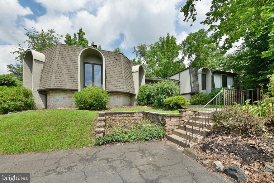This house has a unique architectural design with features suggesting a mid-century modern aesthetic. The structure is characterized by its unusual roofline and curved elements, particularly noticeable in the sections with rounded roof extensions over some windows. The exterior uses a mix of materials, with a stone or textured facade and a lighter stucco or siding underneath. There’s a prominent entryway with wide double doors. The landscaping around the house is simple, with shrubs and a grassy lawn. Overall, the house has a distinctive and somewhat retro appeal. Ready for you to update to your own personal touches.