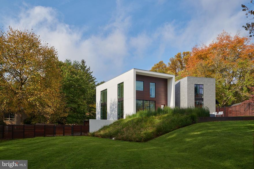 Nestled amidst the verdant bends and winding hills of Chevy Chase, 9119 Levelle Drive is a
textbook showcase of cutting edge modernism.  Designed in 2021 by the acclaimed Robert M.
Gurney, FAIA and built by the renowned Peterson and Collins Builders, the extraordinary skill and craftsmanship is evident throughout and unparalleled.  This singular home is yet another award-winning example of geometry, brilliant manipulation of light, artistic use of glass, and conformation with its natural surroundings. Prominently sited on a coveted lot of just under an acre, towards the end of a no-through road, the approaching driveway opens into a rectangular motor court. Two likewise rectangular volumes are situated in a narrow ‘V’ configuration, connected by a glass bridge housing the front door. A transverse
gallery introduces the fully open concept main level by its informal spaces first. The chef’s
kitchen anchors said space, blending design and function with floor-to-ceiling minimalist, light
wood cabinetry concealing a comprehensive suite of commercial-grade appliances. An
extraordinary oversized center island, clad in light stone slabs with integrated seating and
waterfall edges, spans the entire length of the room. A flooring change from wood to tile, both
outfitted with radiant heat, signifies the transition into the adjacent light-filled breakfast area. A
generously proportioned, continuous living and dining area comprises the majority of the main
level. The latter is backed by full-height panes of glass, letting in tranquil wooded views. The
former, anchored by an abstract half-wood half-mosaic fireplace wall, interacts with said views
via a wall of glass which can be opened to blur the relationship between indoor and outdoor
living. The central staircase, artfully crafted from wood and glass, is both a showstopper and a
structural feat, which connects all levels of the home. The upper level contains private quarters,
including a stunning primary suite resting amidst the treetops. It features generous closet space
with custom built-ins, and the ubiquitous spa-inspired bath with a soothing ambiance attributed
to neutral woods and large panes of glass, as well as all the usual accoutrements. Secondary
bedrooms are well-proportioned, luxuriously finished, and arranged so as to maximize privacy.
Across the glass bridge is the second volume, clad in dark ground face block to contrast the
main volume’s white stucco. On the ground level, it houses an oversized garage with a frosted
glass door as well as a cozy living space. Upstairs is a fully self-sufficient apartment. The
kitchen mirrors that of the main residence, with full-size SubZero and Viking appliances, and a
peninsula island overlooking wooded views beyond the adjacent living area. A private bedroom
featuring a large walk-in closet, as well as a luxurious fully-tiled bath complete the unit.
Enclosed by full fencing constructed of mahogany and steel, the rear lawn comprises terraced
outdoor living spaces designed to engage with the lot’s topography. They include dual-level
patios with a fire pit, ideal for al fresco entertaining, dining, or lounging on a crisp autumn
evening. Further beyond, private rolling hills are surrounded by total privacy. Convenient to the
shopping, dining, and entertainment of both downtown Bethesda and Chevy Chase, as well as
the Nature Conservatory and trails along Beach Drive and major commuter routes, the location
is enviable. Not to be replicated in its architectural significance, 9119 Levelle Drive is a true one-
of-a-kind offering.