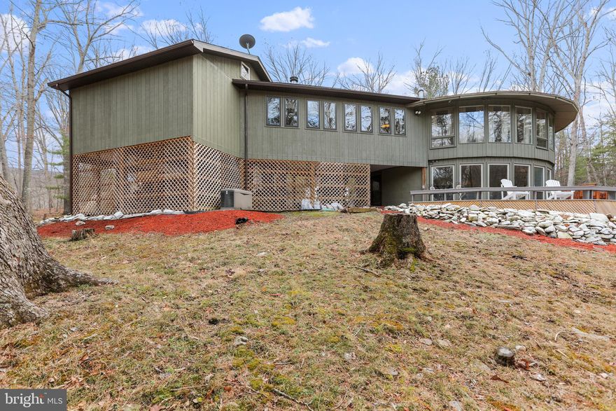 Live in the treetops from this unique River Ridge circular home situated on 6.79 acres . Spacious with three distinctive living areas, it is perfect for full time home,  weekend retreat or an Air BnB.   Delight in the first of two primary suites separated from the main house by a family room breezeway replete with wood stove, making a perfect niche for relaxing or reading on cool days or evening.   This private suite features a sitting area, a large closet, a full bath with dual sinks, jaccuzi tub as well as a separate shower  and a screened-in balcony. Upon entering the main building, bask in the expansive open floor plan with a gourmet kitchen full of new stainless steel appliances, a dining room that comfortably seats eight-plus and is perfectly suited for grand gatherings with space to extend into the casual living room. The second primary bedroom includes an ensuite bathroom, linen closet, built-in bookshelves and two double-doored closets. The spiral staircase leads to a light-filled family room surrounded by windows, built-in bookshelves, and a bar. The lower-level third bedroom offers a quiet respite for weary travelers. An additional separate room currently used as a fitness area can be converted to an office or a secluded den for afternoon naps. Enjoy BBQs and cocktail hour with guests on the massive deck with sounds of the wet weather stream cascading through the property.   Whole house generator conveys and radon system installed.  Plenty of parking, on-site laundry room, and outpost shed for storing bikes, all-terrain vehicles, landscape & garden equipment or tubes for floating the river.   Located in the locked gate community of River Ridge and 1000 =/- feet from the pristine community park with stone BBQ's, picninc tables, storage racks for kayaks/canoes, access to the Cacapon River for floating in a canoe, kayak or tube and bathroom facility on site.