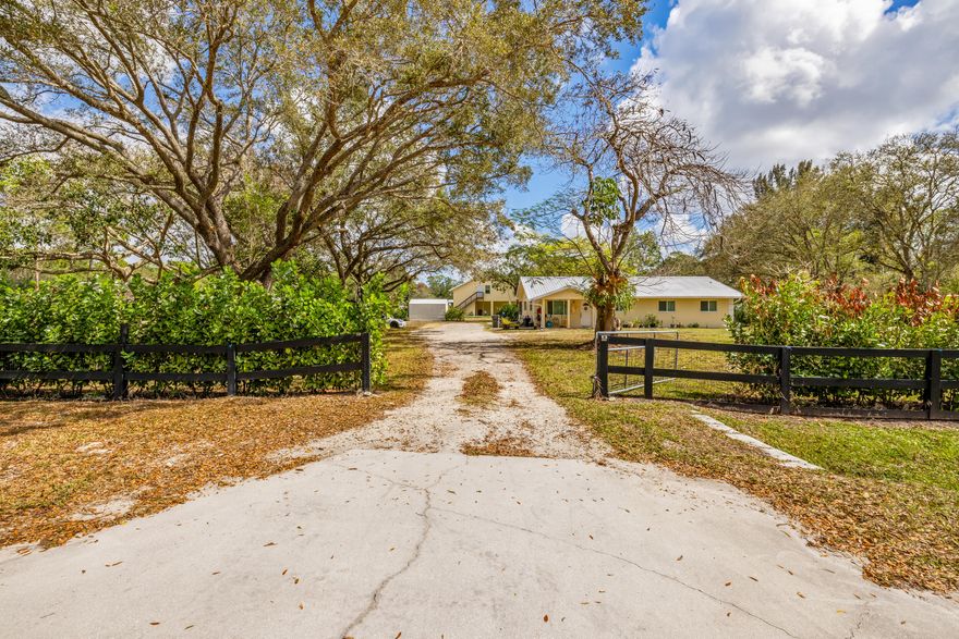 Set on 2.5 fully fenced acres in Jupiter Farms, this versatile property features a 3BR/2BA main home and a substantial secondary structure ideal for additional living space, hobbies, or income potential. Main residence offers over 1,700 sqft with tile flooring throughout, a spacious living area, and updated kitchen and bathrooms. The primary suite includes an ensuite bath with cabana access. Additional highlights include a large utility/laundry room, impact windows, a wired security system, and a covered screened patio perfect for outdoor living. Built in 2015, the secondary structure features a 544 sqft one-bedroom apartment, a 1,242 sqft air-conditioned garage with roll-up door and epoxy flooring, and 192 sqft of storage, all with a separate entrance. Powered by a 150-gallon propane tank