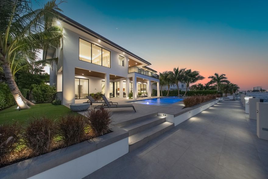 A modern masterpiece sited on the main intracoastal offering unrestricted saltwater and seamless direct ocean access. This estate seamlessly blends sleek architectural design, luxurious amenities, and cutting-edge technology to offer an exceptional South Florida lifestyle. Nestled minutes from Hillsboro and Boca Inlets, this waterfront home is ideal for yachting enthusiasts with its 91-foot yacht dock complete with bumpers, cleats, Shore Power Pedestal, and water supply, all secured by robust concrete pilings and evening lighting. The home's custom architectural design is captivating, combining tropical landscaping with expansive 10-foot PGT impact glass doors that welcome natural light while connecting indoor and outdoor spaces. An entertainer's paradise, the exterior offers a spacious loggia with a summer kitchen, a gas-powered concrete fire bowl for ambiance, and a saltwater pool and spa with sun shelves, LED bubblers, a spillway, and powerful jets all controlled by the smart home automation system. The second-story covered balcony is thoughtfully equipped with cable railings, water spigots, and a smart home interface. Inside, 12-foot ceilings enhance the sense of grandeur, while panoramic water views set the tone for the open floor plan. Sophisticated finishes like European oak engineered hardwood and expansive 48x48 porcelain tiles grace the living spaces, anchored by a water vapor LED-lit fireplace in the formal living area. A custom LED-lit staircase with glass railings and wood capping serves as a stunning architectural feature in the bright, welcoming foyer. The gourmet kitchen is a culinary dream, complete with leathered Taj Mahal quartz countertops, and high-end Monogram and Bosch appliances, including dual built-in refrigerators, a Bosch espresso machine, and a Thermador wine fridge. A jumbo-sized island provides ample seating and prep space, while a central vacuum with toe-kick features keeps the kitchen spotless. The butler's pantry, trimless recessed lighting, and dedicated office space with glass doors and custom cabinetry add both functionality and style. The primary suite is a sanctuary, boasting waterway views from a private terrace, a custom breakfast bar, his-and-her vanities, an elegant makeup vanity, and expansive walk-in closets. The oversized shower, designed as a spa-like retreat, includes dual rain heads, body jets, and floating benches. A mother-in-law suite on the main floor offers privacy and comfort, with easy access to the pool, dock, and outdoor shower via a cabana bath. Equipped with advanced smart home and security systems, the property features a Ring doorbell, four CCTV cameras, motion-sensor flood lights, and comprehensive automation that controls lighting, climate, pool, and security. Energy-efficient features include Low-E glass, tankless water heaters, R-20 foam insulation, gas lines for essential appliances, and a generator-ready setup for added reliability. A two-zone 17 SEER HVAC system ensures optimal temperature control, while a 600 AMP underground electrical service and Cat-6 wiring throughout the home keep it future-ready.  Adding convenience, dual laundry rooms, a mudroom with custom storage, and an oversized three-car garage with car lift compatibility, utility sink, central vacuum, and side-mounted motor provide ample functionality. Thoughtfully designed down to the last detail, a private waterfront oasis offering elegance and cutting-edge technology, for a truly luxurious lifestyle.