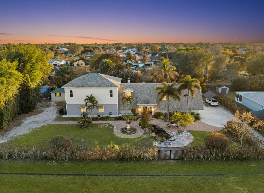 This isn't standard Port Saint Lucie. This is a rare compound-style estate on a fully fenced double lot with architectural presence, outdoor luxury, and the kind of flexibility that doesn't come around often. Offering 5 bedrooms, 3 bathrooms, and over 4,300 square feet of total living space, this home delivers true flexibility with three gated entrances and room for your boat, RV, and recreational vehicles, all without HOA restrictions. Nearly half the home is defined by a dramatic, steep-gabled roofline, an architectural statement rarely seen in typical suburban builds. The layout naturally supports multi-generational living, home offices, creative studios, or future income potential. Expansive yet intentional, the layout allows everyone to live, gather, and retreat with ease. Inside, the main living area makes an immediate impression. A soaring great room rises beneath vaulted ceilings, wrapped by a loft and anchored by a dramatic, floor-to-ceiling custom library wall. An oversized chandelier adds scale, while a wood-burning stone fireplace grounds the space with warmth and texture. The kitchen features white shaker cabinetry, granite countertops, extended peninsula seating, butcher block accents, and skylights that bathe the room in natural light. Outdoors, the experience continues. A brick paver courtyard centers around a serene koi pond with stone. The turf-lined pergola, complete with a built-in fire pit and motorized retractable awning, creates a covered entertaining space designed for year-round enjoyment. An electric-ready workshop or storage area adds practical function, while mature palms and bamboo privacy screening frame the property in lush seclusion. Homes of this scale and character rarely become available. This is a property that blends land, privacy, and architectural identity offering the freedom to host, expand, create, and live without compromise.