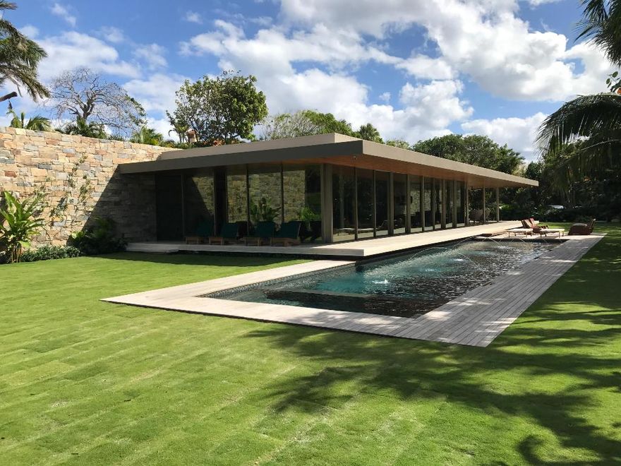 A stunning modern estate set on a rare, oversized 40,000± square foot lot, this exceptional one-story residence is a masterwork by award-winning modernist architect Marcio Kogan of Studio mk27, conceived to blur the boundaries between indoor and outdoor living. The home offers four bedrooms, five full baths, and two powder rooms, arranged within a thoughtfully curated floor plan that balances openness with privacy. Expansive floor-to-ceiling Vitrocsa glass walls flood the interiors with natural light and allow the living room to transform effortlessly into an open-air pavilion, overlooking lush, manicured gardens. Architectural details are both refined and intentional: teak rods line the ceilings, while travertine flooring extends seamlessly from interior to exterior, visually and physically uniting the spaces. The indoor living room, fully enclosed in glass, opens to tranquil garden views, while the outdoor living room is anchored by a dramatic metal fireplace suspended from the ceiling, creating a striking focal point.
A sculptural Virginia flagstone wall artfully separates the social areas from the private wing, which includes the bedrooms, kitchen, office, and media room. The primary suite is a serene retreat, featuring an oversized walk-in closet, dual baths, and peaceful garden vistas.
The exterior spaces are equally compelling. Loggias, a resort-style pool area, and lush tropical landscaping create a private outdoor oasis, enhanced by a dramatic living wall that serves as both an artistic statement and a unifying design element between inside and out.
The residence is further elevated by impeccable interior design by Diana Radomysler and landscape architecture by Keith Williams of Nievera Williams Design. Recognized for its architectural significance, the home is the Winner of the Preservation Society's 2020 Schuler Award.
This is a truly one-of-a-kind estate; a residence that is as artistically compelling as it is functionally sophisticated.