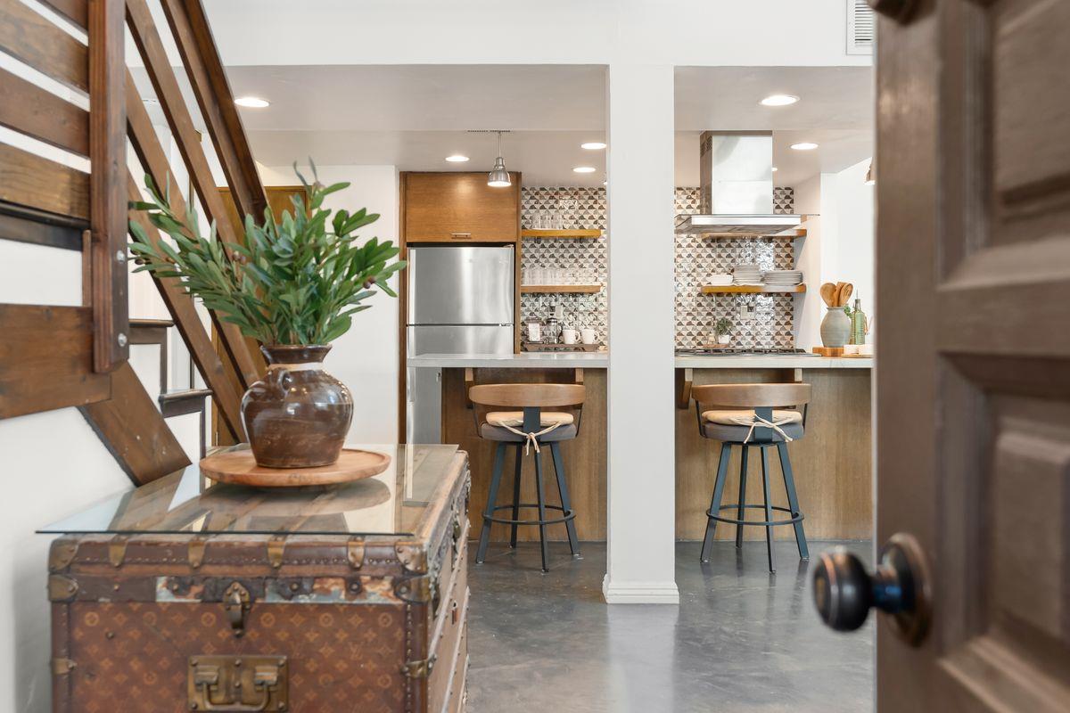 Condo kitchen with patterned tile backsplash, open wood shelves, and two bar stools; trunk and staircase in foreground.