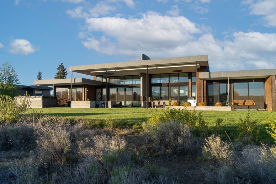 Located on one of Tetherow's premier lots, this architectural statement celebrates the beauty of the high desert with panoramic views of the Cascade Mountains. The great room commands attention with soaring, curved 14-foot ceilings and a 46-foot expanse of glass that frames the western skyline. Crisp lines, radiant heated floors, and a newly crafted stone accent wall reflect a modern, yet timeless aesthetic, while an indoor/outdoor fireplace connects the soul of the home to the landscape beyond. Designed for both entertaining and private retreat, the home offers four luxurious en-suite bedrooms, a fitness studio, a dedicated office, and a rooftop terrace ideal for sunset gatherings. The reimagined primary suite rivals any five-star resort, offering a refined sanctuary of texture, warmth, and contemporary elegance. Outside, an expansive patio with a fire pit, and an enclosed courtyard elevate the outdoor living experience, ensuring every season is enjoyed to its fullest.