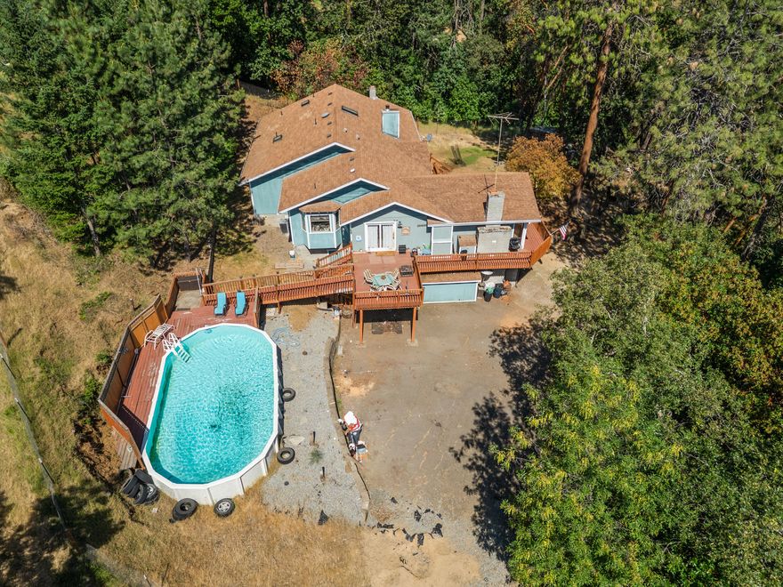 Enjoy privacy and mountain views on this serene 3.12-acre property featuring an above-ground pool with a deck. Natural light floods the interior through large windows and skylights. The kitchen is ideal for entertaining with granite counters, a center island stove, and range hood. Relax in the open-concept living room or step onto the wraparound deck for fresh air and panoramic views. The spacious home offers a large primary suite with fireplace, vaulted wood ceilings, and a jetted tub. The home boasts 3 bedroom plus 2 bonus rooms currently being used as bedrooms. A dirt road behind the home leads to a flat area—perfect for a shop, RV pad, or outdoor retreat space. A peaceful getaway just minutes from town.