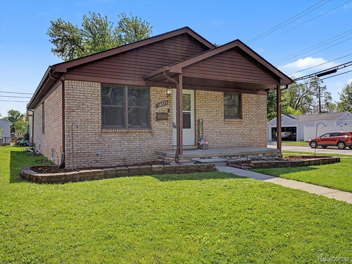 Brick ranch home featuring a covered front porch, concrete walkway, established lawn, and a decorative stone planter