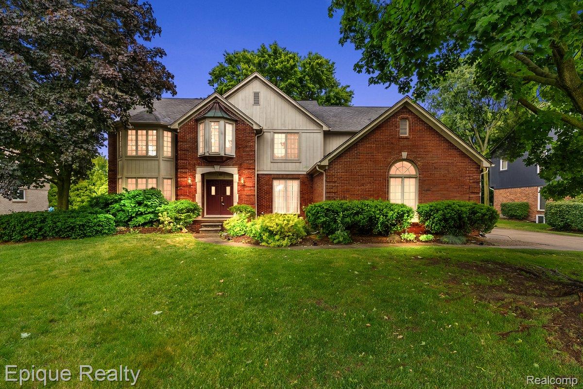 Brick and siding exterior featuring a prominent bay window, gabled roofline, mature trees, manicured shrubs, and a well-maintained lawn
