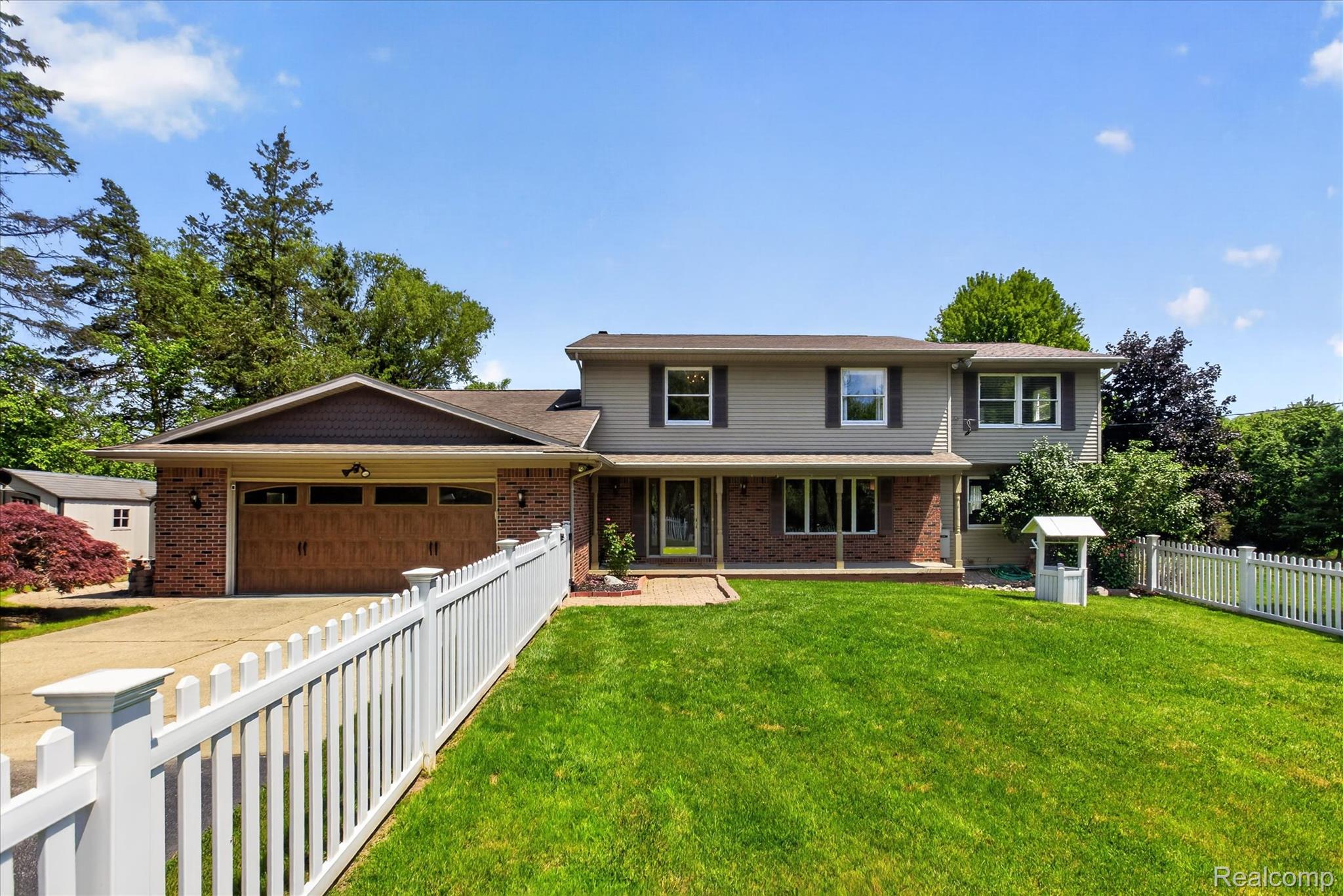 Two-story residence featuring a brick and siding exterior with dark brown shutters, an attached two-car garage with a wood-finish door, a manicured front lawn, and a white picket fence