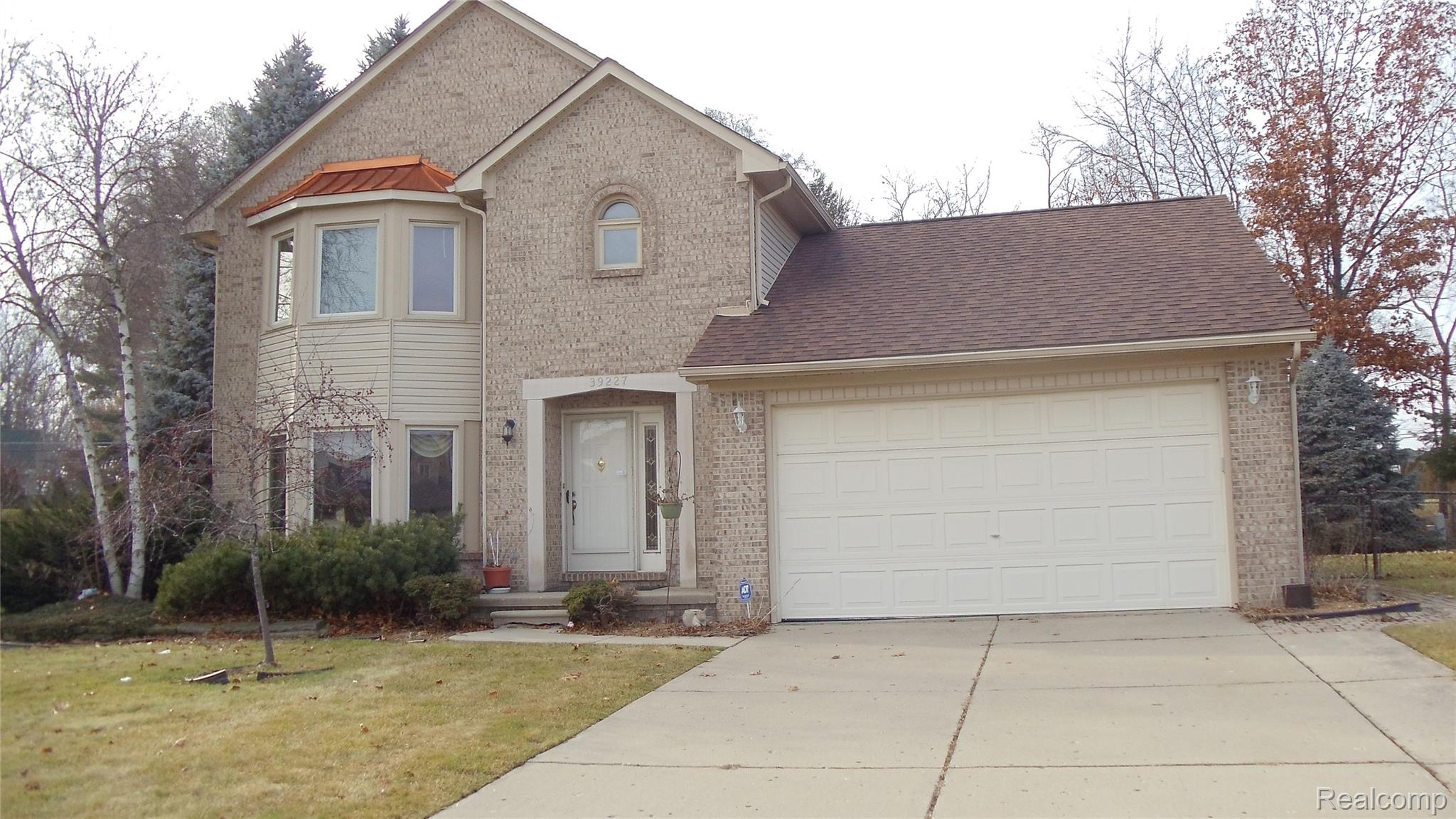 Brick exterior featuring a two-car garage, bay window with metal roof accent, arched window detail, and mature landscaping