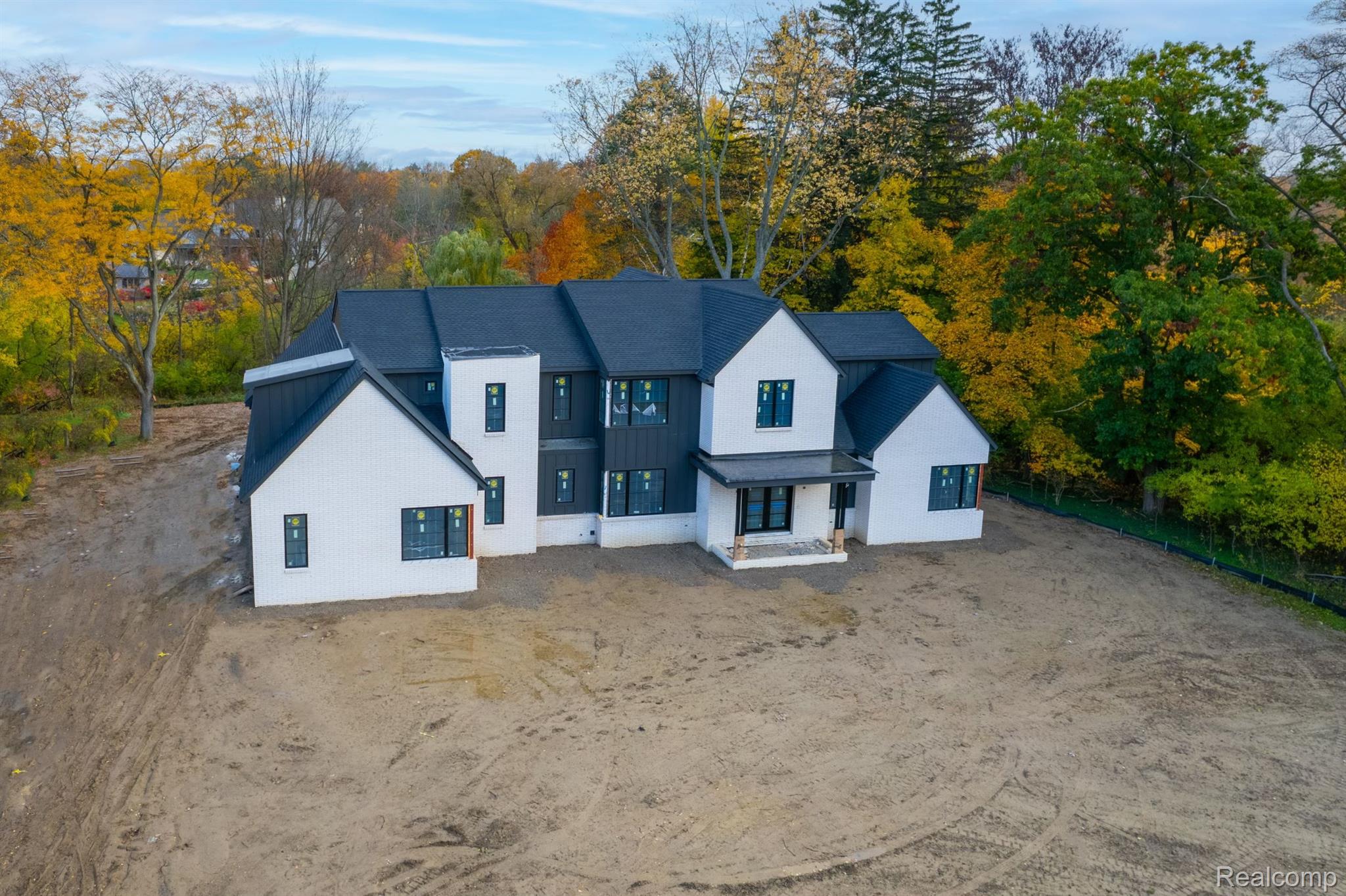 Modern two-story house under construction, with white brick, dark siding, and autumn trees behind it.