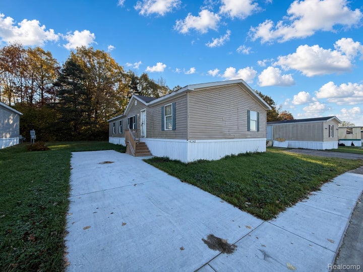 Light tan horizontal siding exterior featuring dark shutter accents, white skirting, a concrete driveway, and wooden…