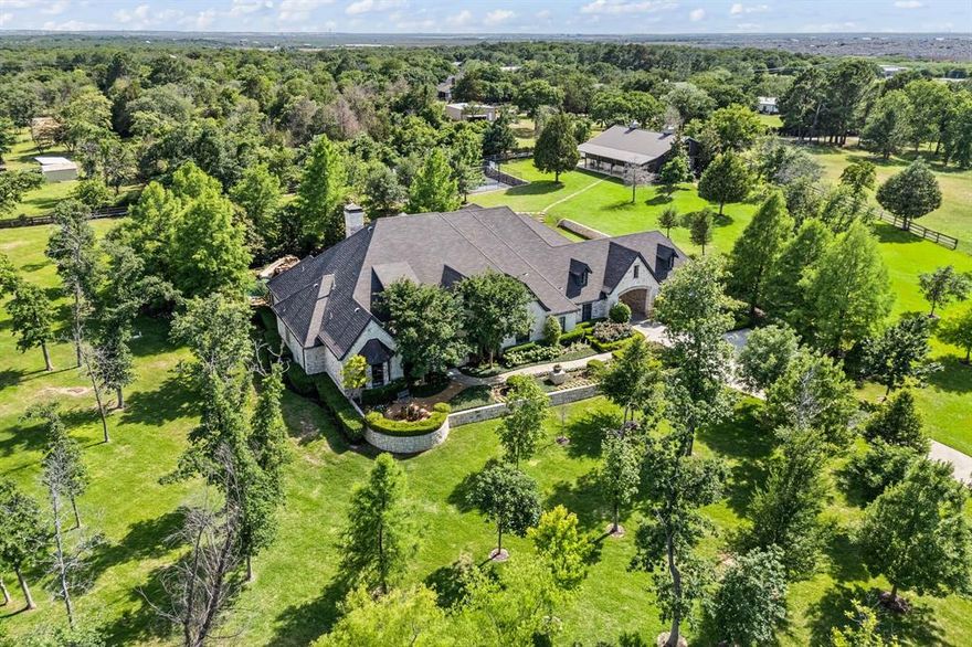 Some homes greet you at the gate and never let go. This one-story custom estate in Argyle does just that- tucked behind a private entrance on over five acres, where the beauty of wide-open land offers a rare blend of quiet country atmosphere, all within the boundaries of highly sought-after Argyle ISD. The drive winds past manicured lawns, as the world slows down, and up to a timeless estate. Step inside soaring beamed ceilings draw your eyes up, while warm wood floors and the crackle of a stone fireplace create a sense of welcome. It’s the kind of space where family moments unfold naturally- where glass doors slide open and the line between indoors and out becomes blurred.
Five spacious bedrooms, each with its own ensuite bath, offer restful corners for everyone. There’s a quiet ease in knowing that comfort and privacy were considered in every inch of the design. This isn’t just a home for everyday living- it’s a place built for gathering, for making memories, for staying a while. Outside, the lifestyle continues to unfold. A deep covered patio beckons with the glow of a second fireplace- ready for a cozy morning coffee or long summer nights with friends. Just beyond, the pool sparkles in the Texas sun, complete with a waterfall, slide, and spa.
Toward the edge of the property, a grand 2,520 square foot barn stands tall. Tucked inside the barn is a guest home with private bath, perfect for in-laws, weekend guests, or even a private office. And just steps away, a full-size tennis court stretches out, ready for early morning sets or evening matches under the stars. A nearby firepit circles up beneath the sky, the kind of spot that naturally draws people together.
This property isn't just about space—though there’s plenty of it. It’s about what that space makes possible. Slow mornings. Lively gatherings. Peaceful evenings. Family.