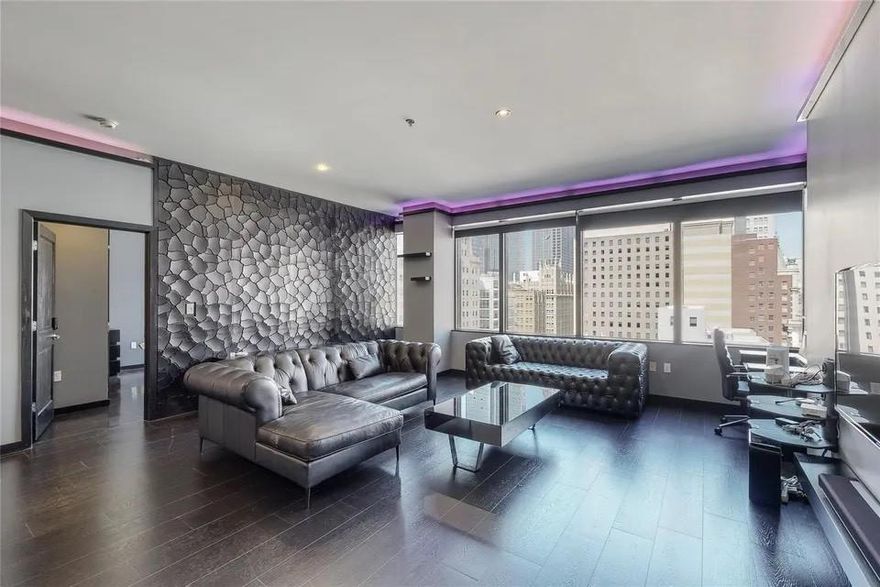 High above the pulse of Downtown Dallas, this 15th-floor residence feels like a private perch in the sky, where morning light spills across dark hardwoods and the city’s architectural silhouettes frame every moment. Step inside and the atmosphere shifts: soft LED cove lighting adds a modern glow, textured feature walls create depth and dimension, and expansive windows draw your eye outward to the skyline that becomes part of the home itself. The main living area opens naturally, giving each space room to breathe. A clean-lined, contemporary kitchen anchors one side with thoughtful cabinetry, stone counters, and stainless-steel appliances, designed for easy movement whether you’re cooking, working, or unwinding. Remote-control shades in every room offer instant calm at the touch of a button. The private bedroom suite feels tucked away from the world, with a custom walk-in closet and an ensuite framed by warm stone tile. A second bedroom adds versatility, an inspiring home office, guest sanctuary, or creative nook, each with access to a balcony that captures the hum of the city from a peaceful distance. Beyond the walls of the unit, the building’s amenities extend the experience: a rooftop pool wrapped in sun and skyline, a full fitness center, a theater room for quiet evenings, lounge spaces for gathering, and a beautifully designed lobby that sets the tone the moment you arrive. Concierge service is available around the clock, offering ease and reassurance with every step. Here, daily life feels elevated, connected to the vibrancy of downtown yet shaped by the comforts of a thoughtfully curated home. A place where modern design, simplicity, and city living meet in effortless rhythm