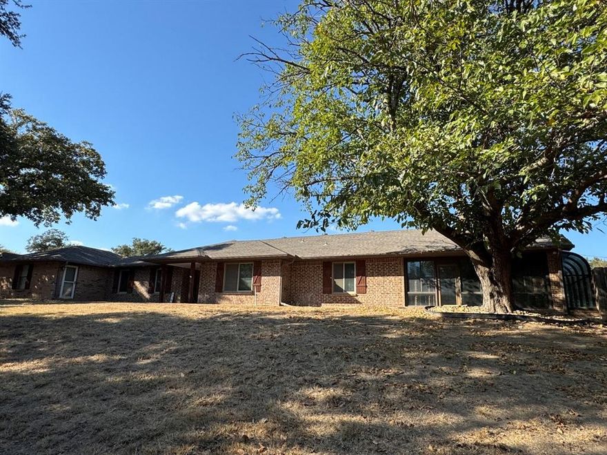 Spacious custom-built single-story brick home located on a scenic .913-acre corner lot in the highly sought-after Aledo ISD! Surrounded by mature oak trees, this versatile property offers 4 bedrooms, 3 full bathrooms, an office, den, formal dining, breakfast area, a 3-car garage, 3-car carport, and a versatile sunroom or game room with a full bar, sink, refrigerator, and poolside access—ideal for entertaining guests or relaxing in style. A private bedroom with a full bathroom is directly accessible from the game room, making it perfect for use as a guest suite, second primary bedroom, or private retreat. The flexible floor plan provides rooms that can easily serve as a 5th bedroom, second living area, or additional office. The chef’s kitchen features a massive granite island, handcrafted cabinetry, farmhouse sink, pot-filler, stainless steel gas range, dishwasher, and window seat.  The living room includes a brick fireplace and built-in cabinetry.  Outdoor living is elevated with a remote-controlled Arcadia motorized louvered patio cover, offering shade or sun on demand. This home blends comfort, flexibility, and function in one of North Texas’ top school districts.  All measurements, square footage, and school information are approximate and should be independently verified. This property is owned by the Secretary of Veterans Affairs and is being sold AS IS.