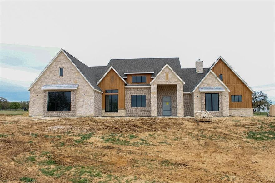 Experience elevated country living in this exceptional new construction home, that blends modern, rustic and classic elements, set on 2 expansive acres. Offering 3,627 square feet of thoughtfully designed space, this 4 bedroom, 3.5 bath residence also includes a dedicated office and a versatile game room or flex space. Designed with entertaining in mind, the open concept layout flows seamlessly into a resort-style outdoor living space. The expansive covered patio features a stunning wood paneled ceiling, a wood burning fireplace, and a sliding glass door that creates a true indoor-outdoor experience. The game room offers direct access to the patio, making it the perfect extension for hosting and gathering. The chef’s kitchen will be both functional and beautifully appointed, complete with a large island, gas cooktop, and designer finishes. A separate prep kitchen adds an elevated level of convenience, featuring a double oven, microwave, and abundant storage, ideal for entertaining while keeping the main kitchen pristine.
The exterior showcases a sophisticated blend of brick, stone, and Douglas Fir accents, complemented by a 3-car garage and side entry driveway for a clean, upscale presence. With premium finishes, intentional design, and ample space to enjoy both indoors and out, this home offers a rare combination of luxury, comfort, and modern Texas living.