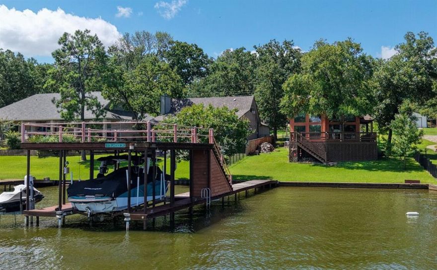 Lodge-style lake home with elevated views and a generous wraparound deck, perfectly positioned in a wide, calm cove with open water views—ideal for swimming, paddleboarding, kayaking, and other water sports. Inside, soaring ceilings, expansive windows, scored concrete floors, and a rustic stone fireplace set the tone for relaxed lake living. The open-concept layout features two bedrooms on the main level and a spacious upstairs primary suite, plus a versatile loft—ideal for extra sleeping space, an office, or game room. The kitchen offers granite countertops, stainless appliances, and custom finishes throughout. The private boathouse includes a party deck, upgraded remote-control lift (designed for a Ski Nautique), and heavy-duty electric swing for a PWC lift. A single-car garage with electric lift adds convenience, and the home is being sold furnished, making it move-in ready. The yard is fully enclosed with wrought iron fencing, and the Hardi Board siding mimics cedar for a rustic look without the upkeep.