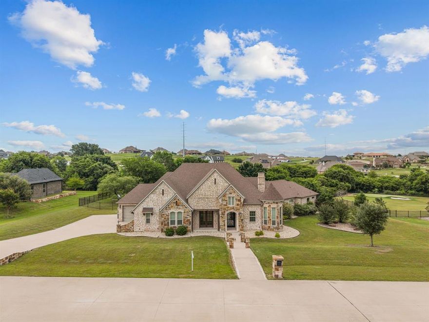 This beautifully designed single story home in the guarded and gated Bella Flora community in Aledo ISD offers 3,972 sq. ft. of thoughtfully crafted living space on 1.28 acres. Interior features include tumbled marble travertine floors, vaulted beamed ceilings, plantation shutters, crown molding, custom lighting, built-ins, and Knotty Alder cabinetry throughout.
The chef’s kitchen is equipped with KitchenAid appliances, double islands, 3cm leathered granite countertops, a six-burner gas cooktop, walk-in pantry, butler’s pantry, and dry bar — ideal for both everyday living and entertaining. Open to the spacious living room with fireplace and built-ins, the kitchen flows seamlessly into the main living areas. A large sliding glass door opens to the backyard retreat featuring a covered patio, built-in grill and bar area, resort-style pool with beach entry and bubblers, grotto waterfall and slide, and a spacious backyard with room for pets and play.
The media room features a barrel ceiling, LED lighting, Epson projector, and surround sound system. The spacious primary suite includes a fireplace, sitting area, and an ensuite bath with marble inlay flooring, separate tub, walkthrough shower, and a fabulous walk-in closet with a hidden storage area.
Three additional ensuite bedrooms provide flexible space for family and guests, including two connected by a Jack-and-Jill bath. Additional highlights include a formal dining room with tray ceiling, custom iron front door, wood beams, and detailed ceiling treatments throughout the home.